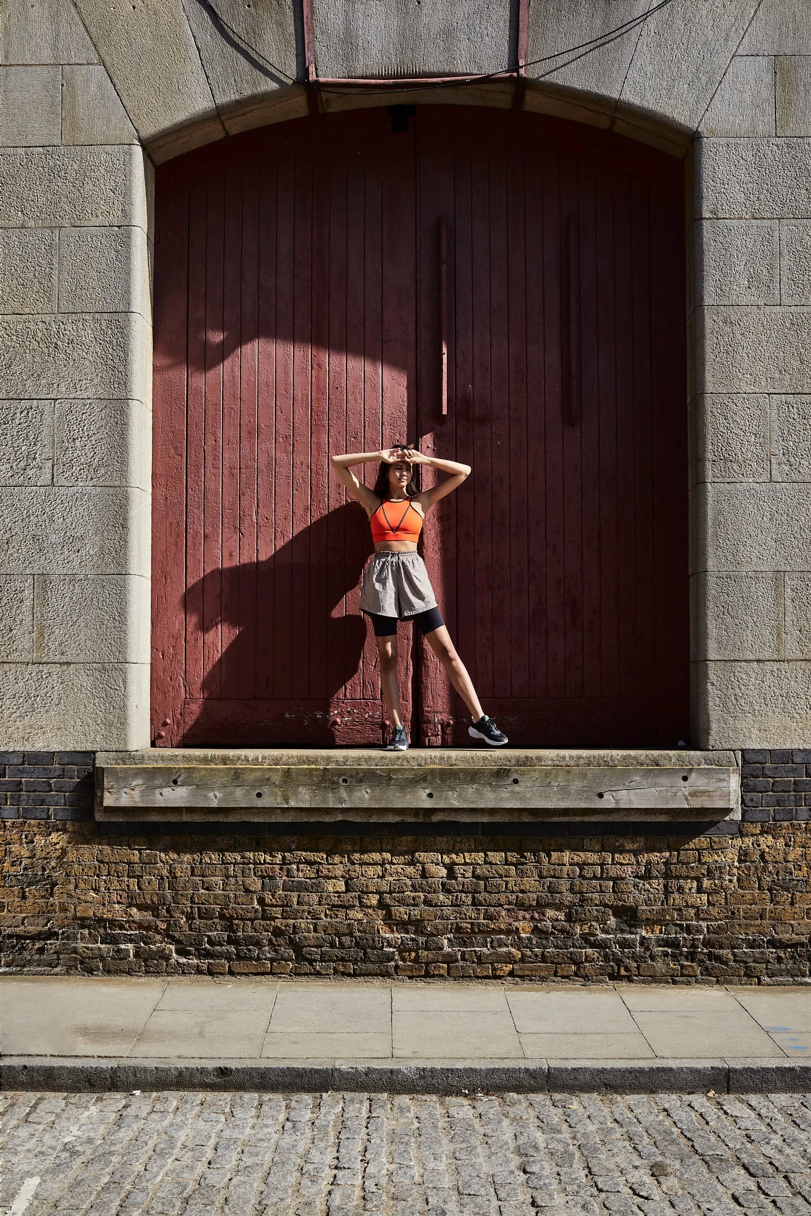 A woman in athletic clothing standing on a small platform in front of a large red wooden door, casting a shadow on the door under sunlight.