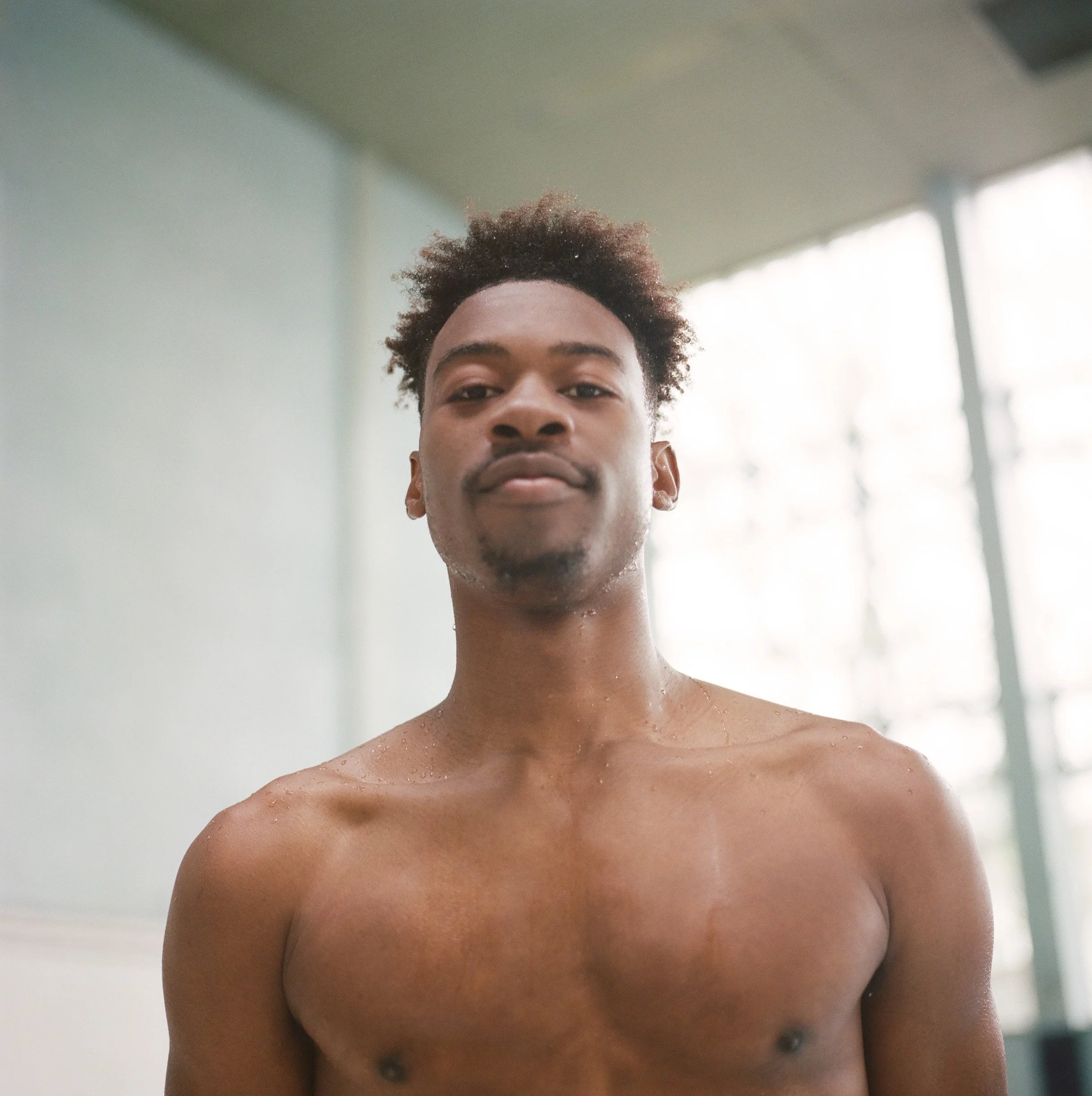 Young man with short curly hair, shirtless, standing indoors near large windows with a blurred background.