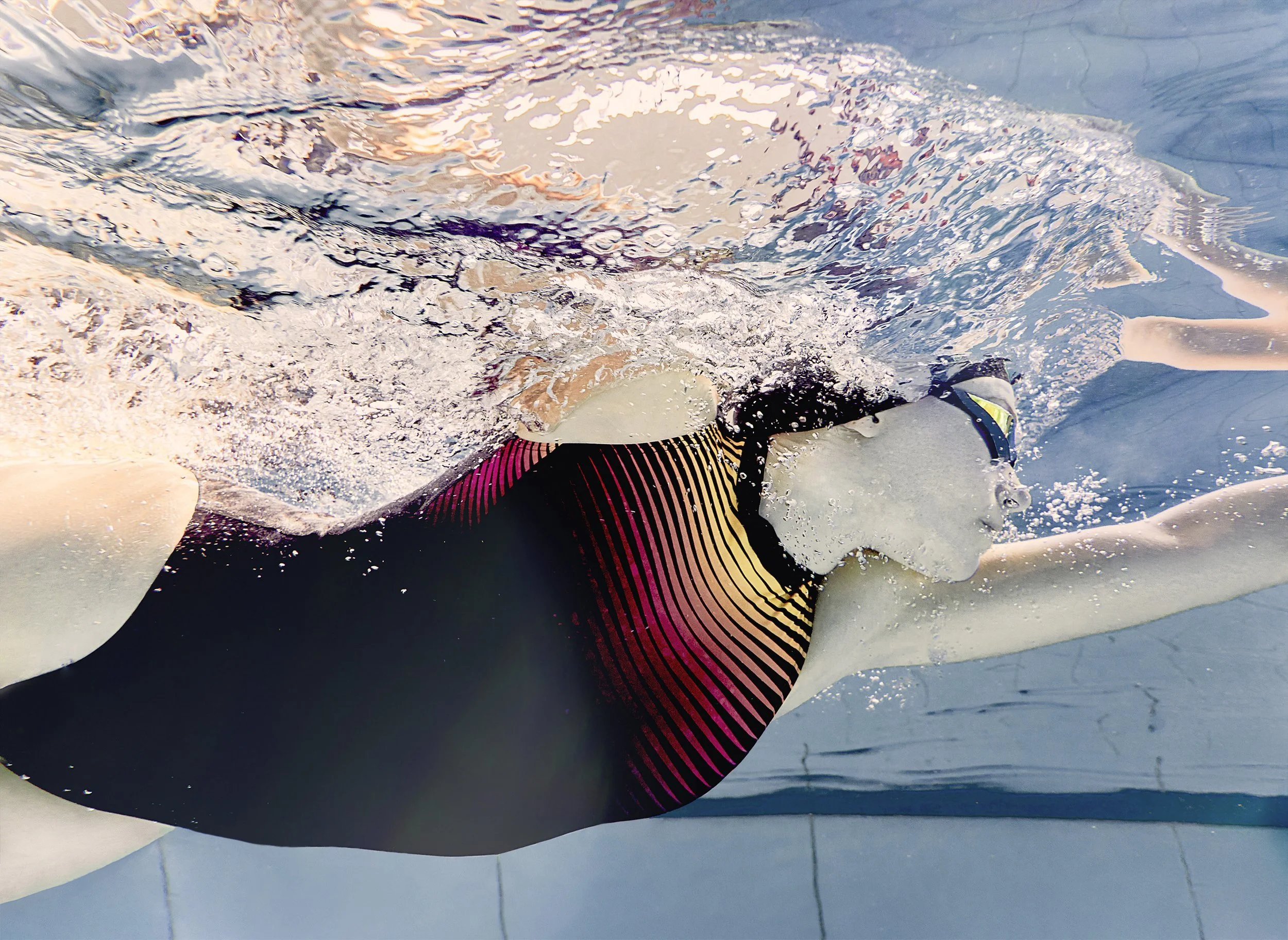 Person swimming underwater in a pool, wearing goggles and a black swimsuit with red and yellow stripes.