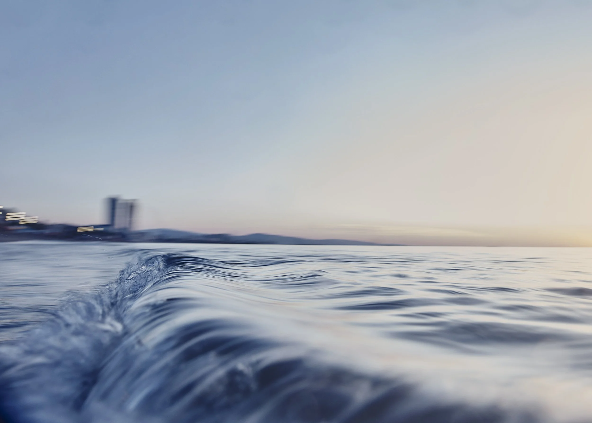 Photo of water with gentle waves, with a cityscape and mountains in the background at sunset.