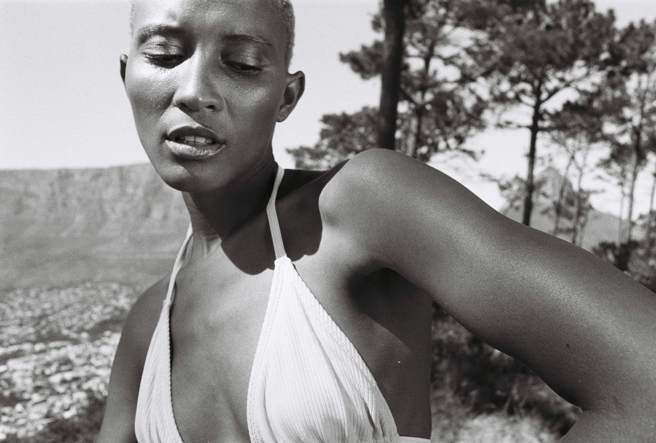 A close-up of a woman with short hair wearing a swimsuit, standing outdoors with trees and mountains in the background, captured in black and white.