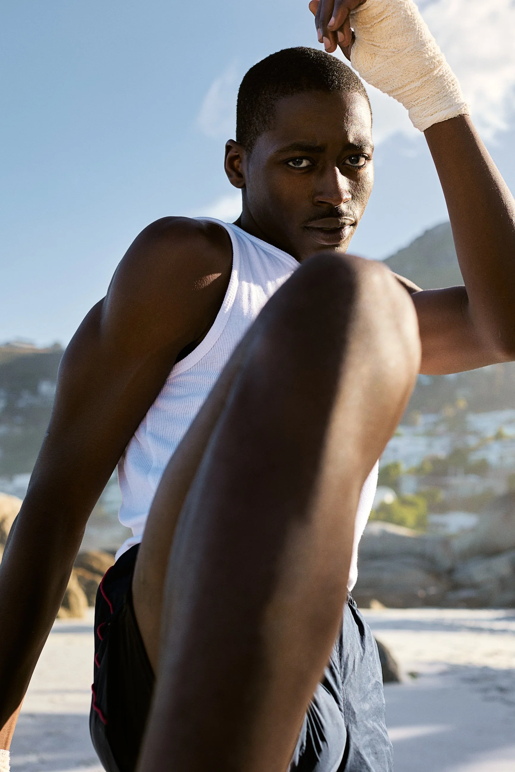 A young man with short hair and dark skin is sitting outdoors on a sunny day, wearing a white sleeveless shirt and dark shorts, with a white wristband on his right wrist, holding his knee with his right hand, against a background of blue sky and rock
