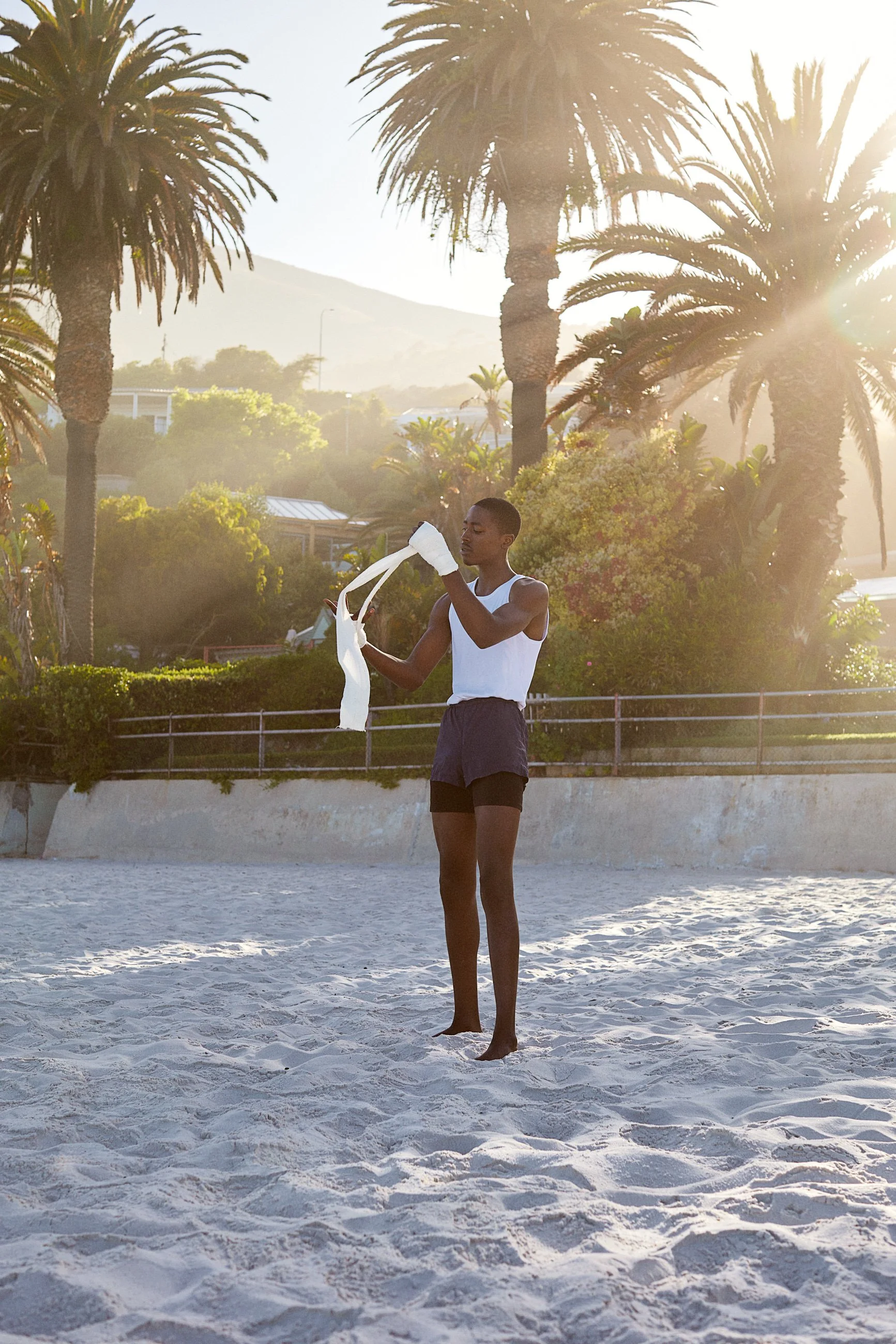 Young person on a sandy beach with palm trees in the background, wrapping a long white ribbon, during sunset.
