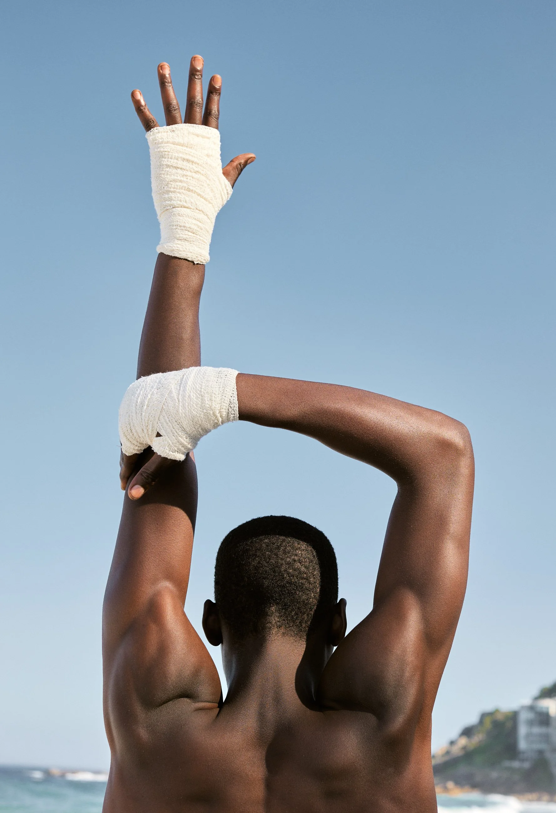 A person with dark skin standing on a beach, raising their right arm with a wrapped wrist and hand, looking towards the sky against a clear blue sky with a distant shoreline and greenery in the background.