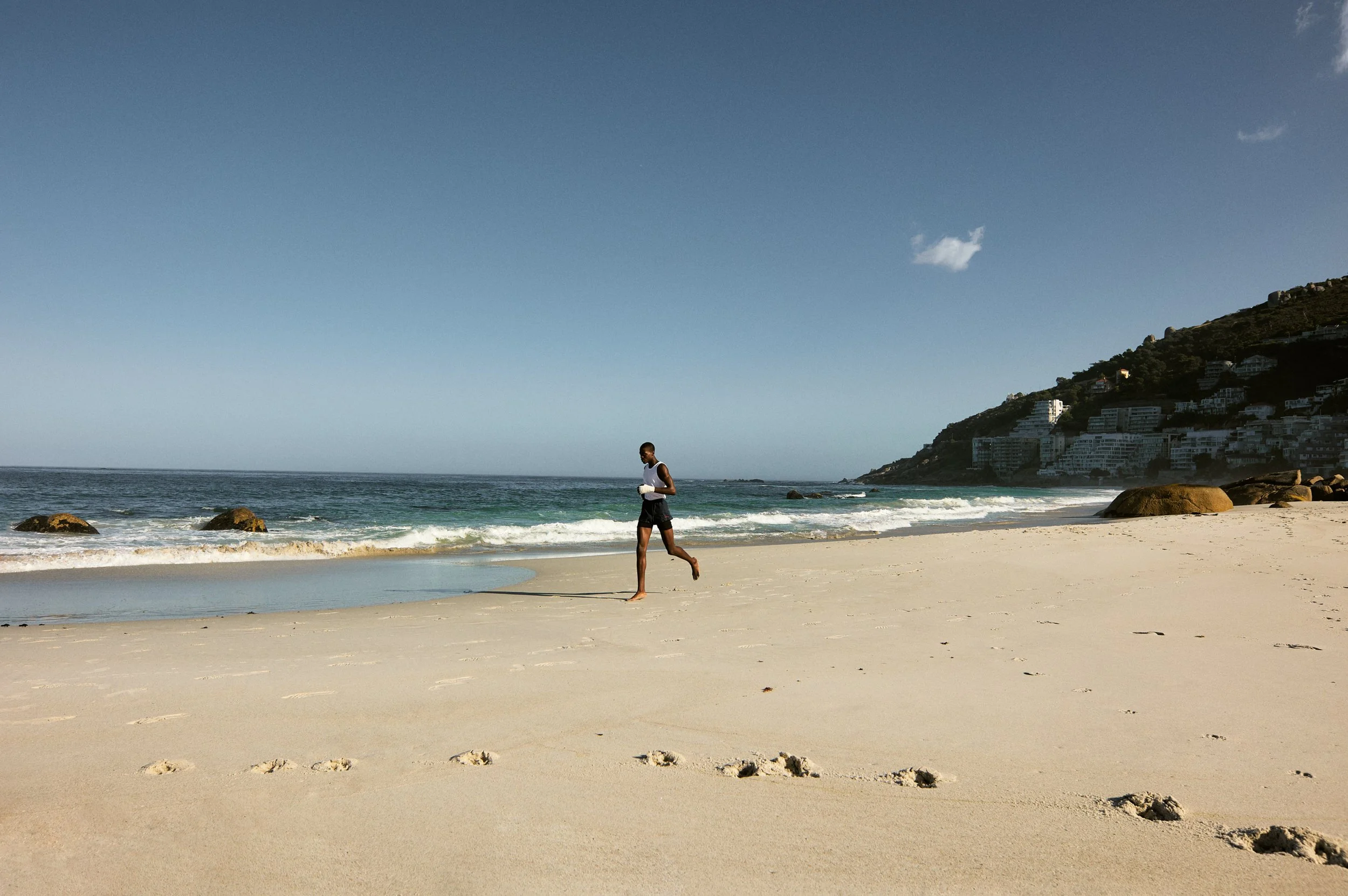 A person running on a sandy beach with footprints, ocean waves, and hillside buildings in the background.