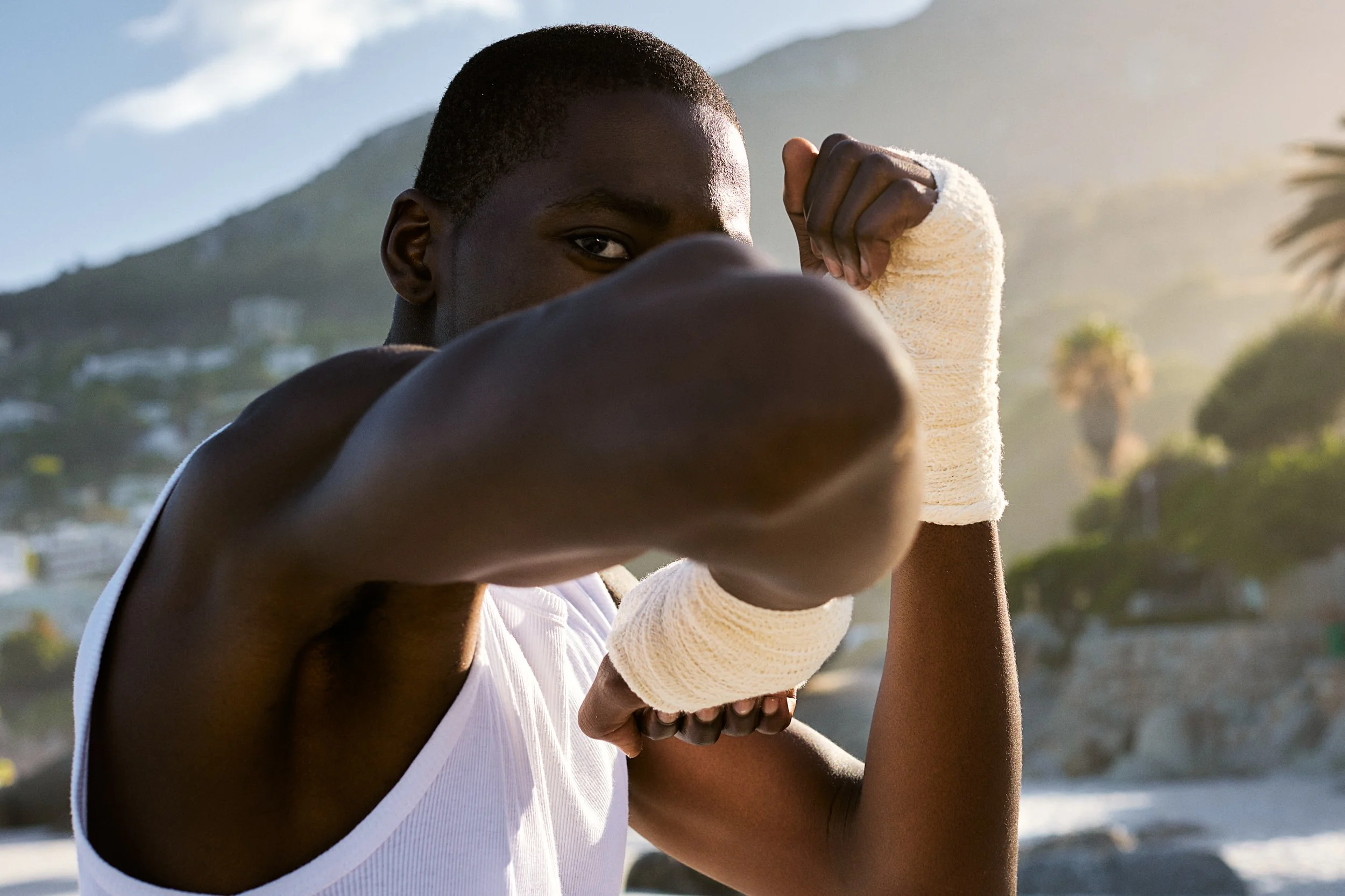 A young man with a caply broken arm in a white cast striking a boxing pose outdoors at a beach, with hills and palm trees in the background.