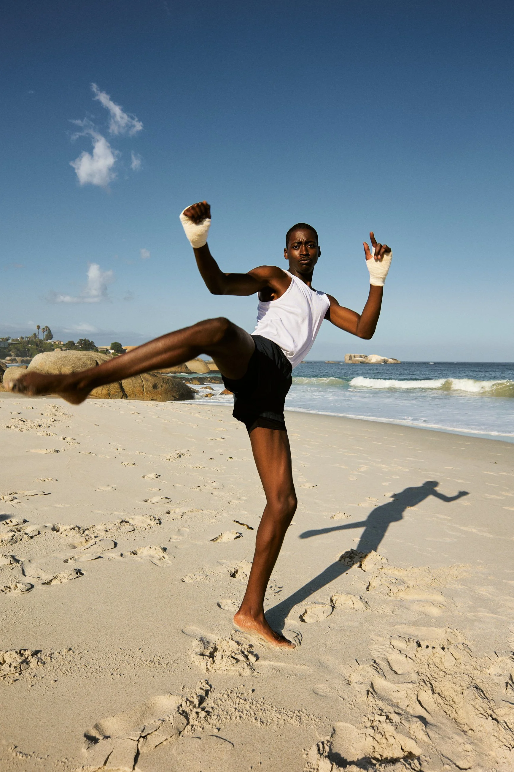 A young man practicing martial arts on a sandy beach with the ocean in the background, wearing a white tank top and black shorts, with boxing wraps on his hands.