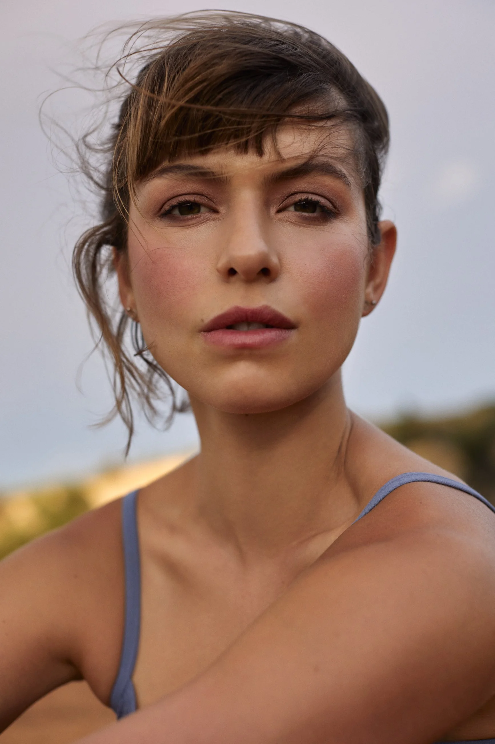 A woman with short brown hair, wearing a blue tank top, looking directly at the camera outdoors during daylight.