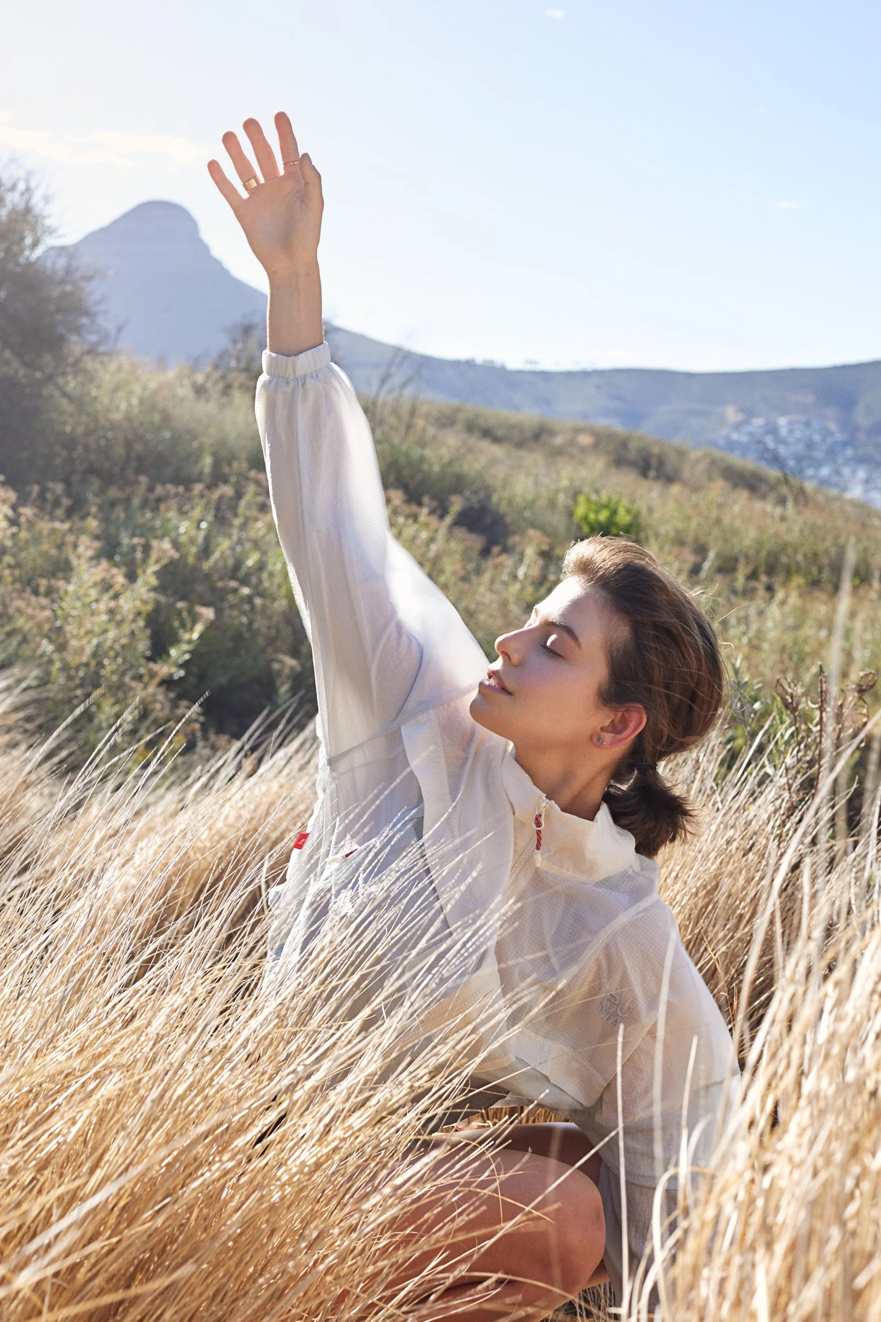 Young woman with brown hair in a braid wearing a cream-colored jacket, kneeling in tall dry grass with mountains in the background, enjoying the sunlight with her eyes closed and one arm raised.