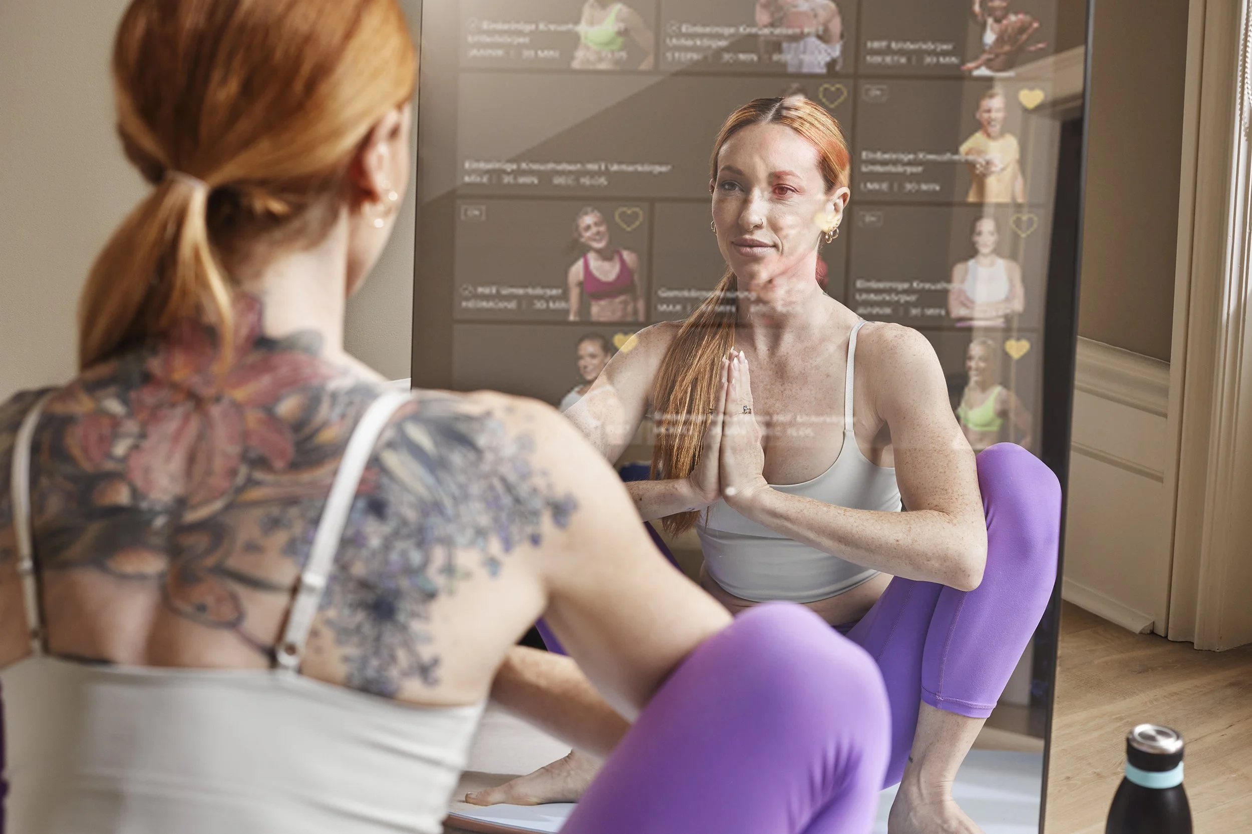 A woman practicing yoga in front of a mirror during a virtual class, with her reflection visible showing her in a yoga pose, wearing purple leggings and a gray top. There is a water bottle on the floor nearby.