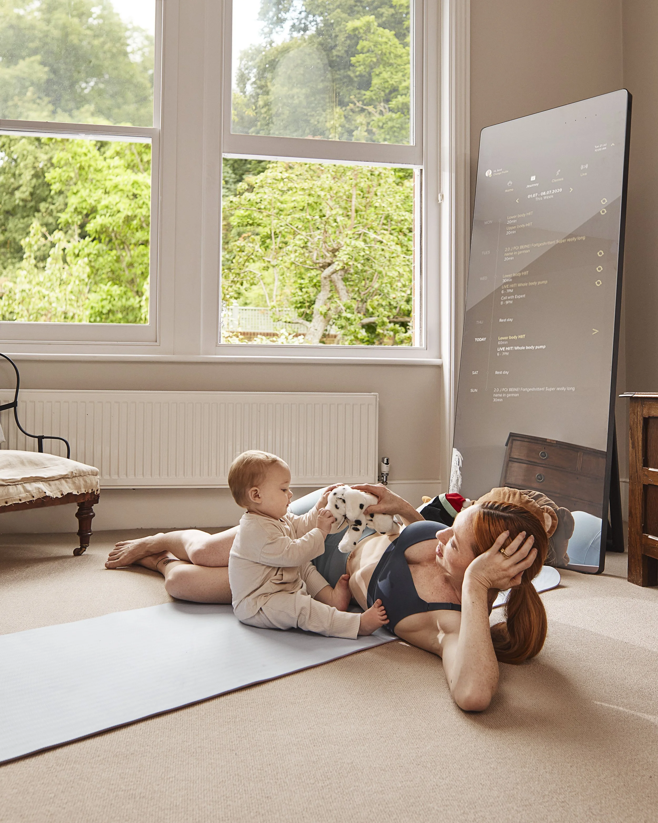 A woman exercising on a yoga mat with a young child playing with a stuffed Dalmatian dog toy beside her in a bright room with large windows showing green outdoor trees.