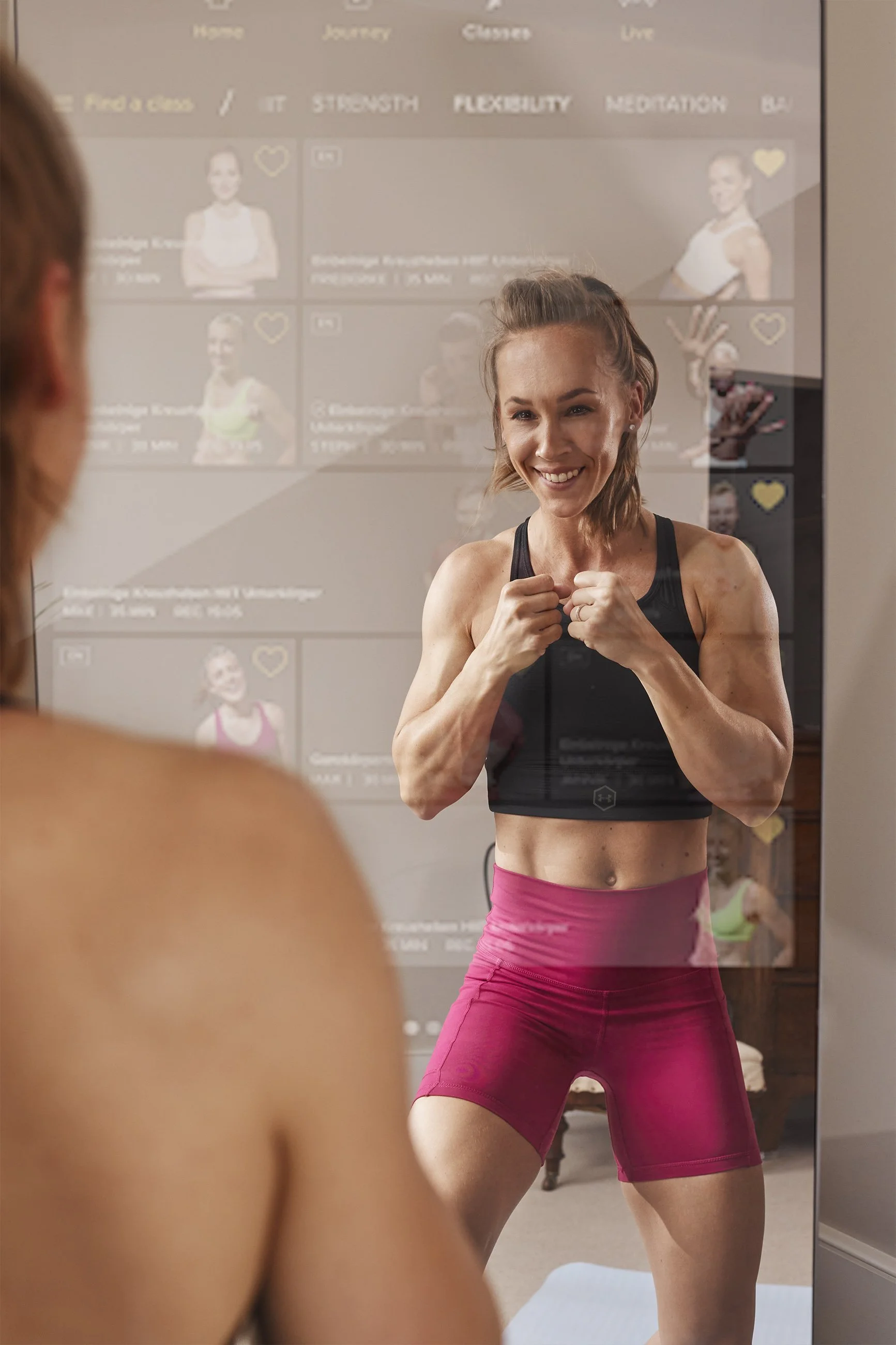 A woman in workout clothes smiling at herself in the mirror, with her arms raised in a victorious pose.