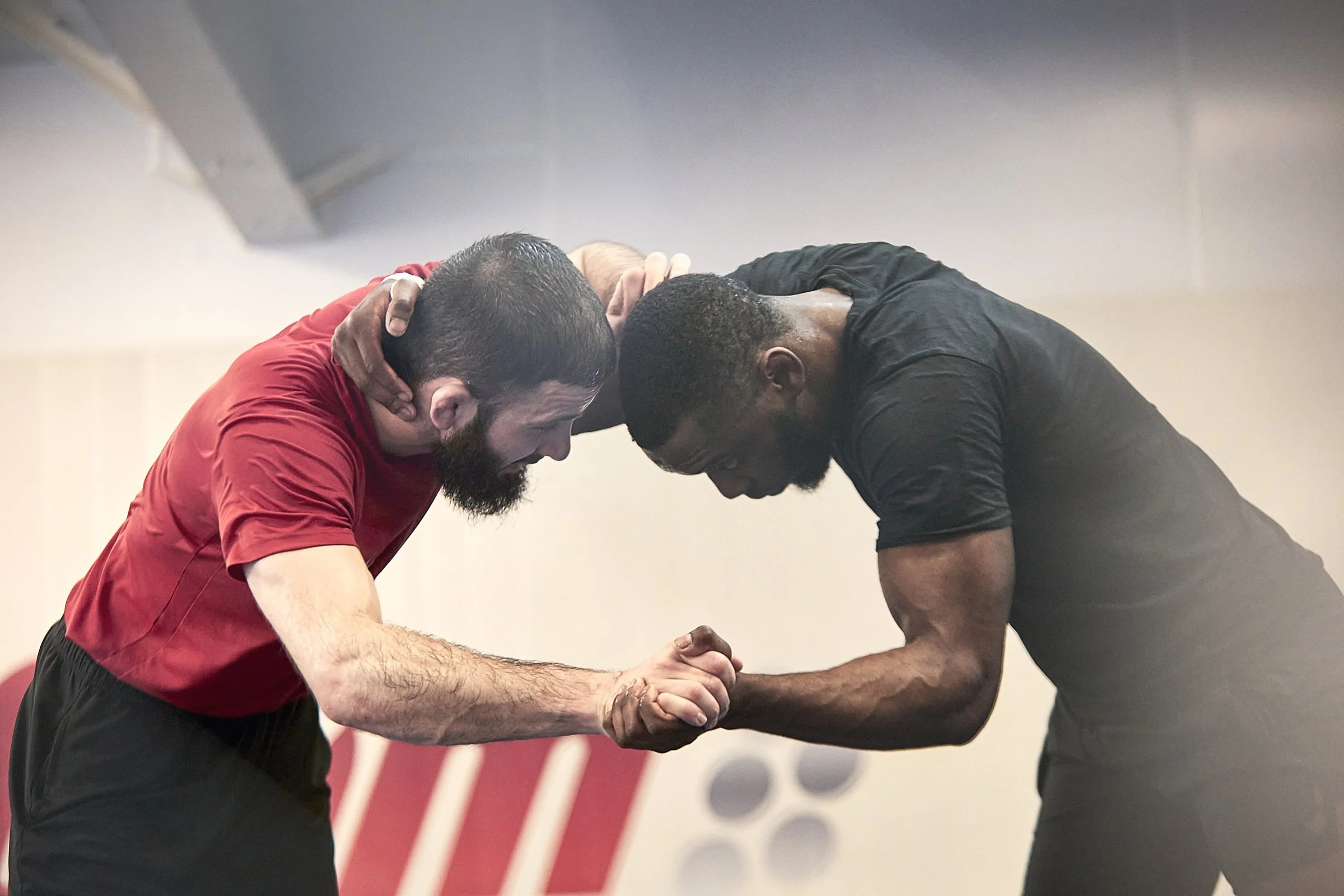 Two men are engaged in an arm wrestling match, leaning towards each other with forearms clasped, in a gym or training area.