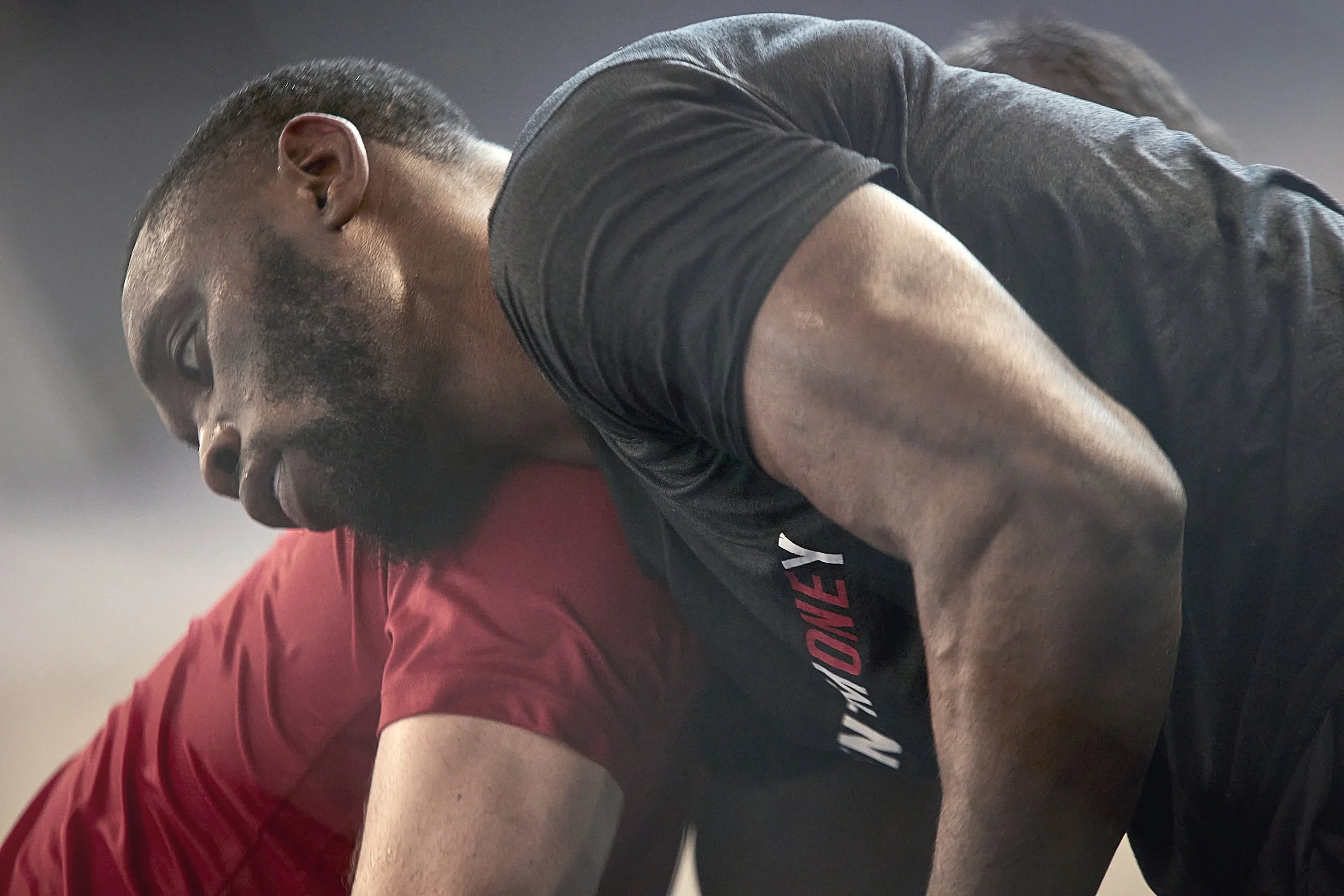 A man with a beard and short hair leaning over, wearing a dark athletic shirt. Another person in a red shirt is partly visible beside him.