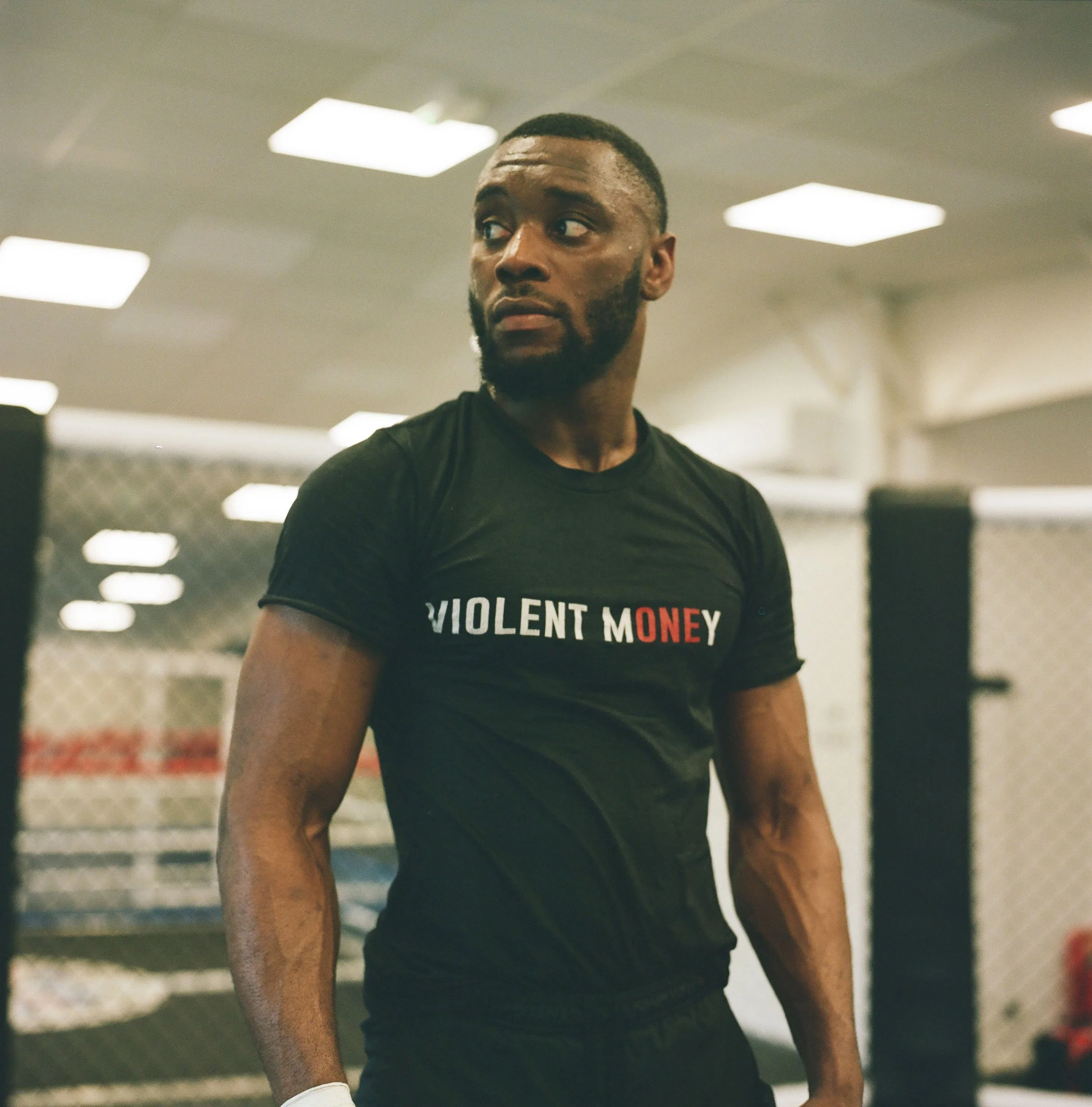 A man in a black T-shirt with the words 'VIOLENT MONEY' printed on it, standing inside a gym with a chain-link fence and workout equipment in the background.