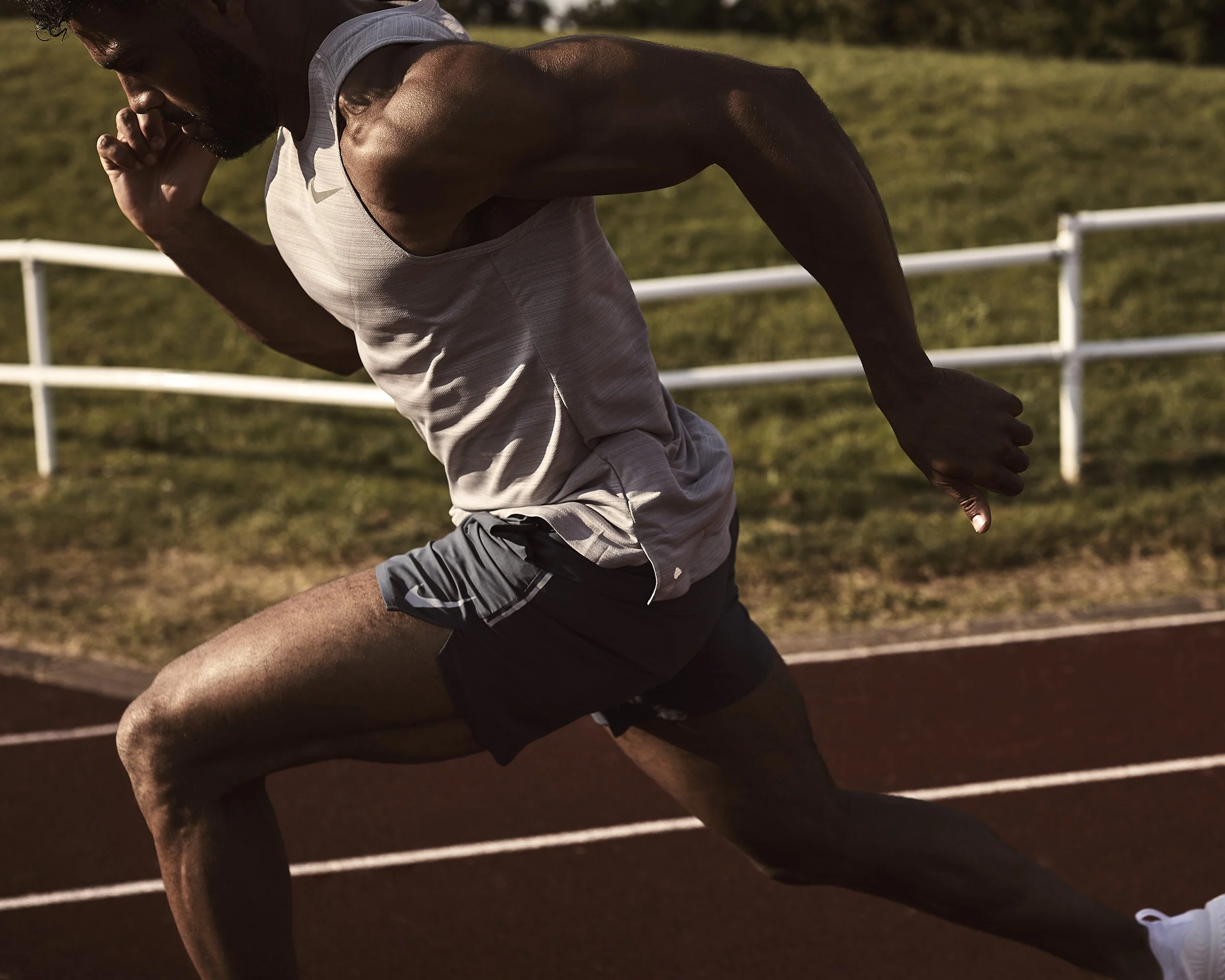 A male athlete in a white tank top and black shorts running on a track outdoors during daytime.