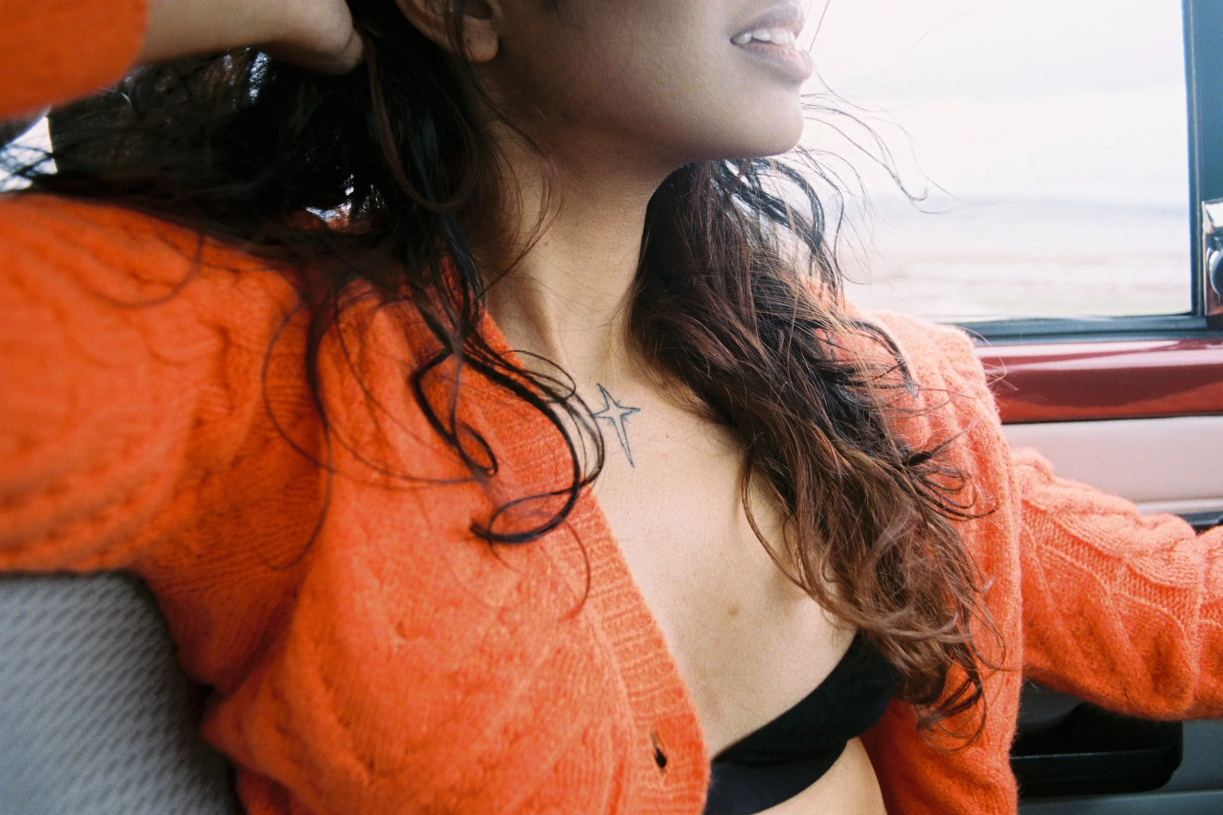 A woman with dark wavy hair wearing an orange sweater and a tattoo of a cross on her chest, sitting in a vehicle, smiling and looking out the window.