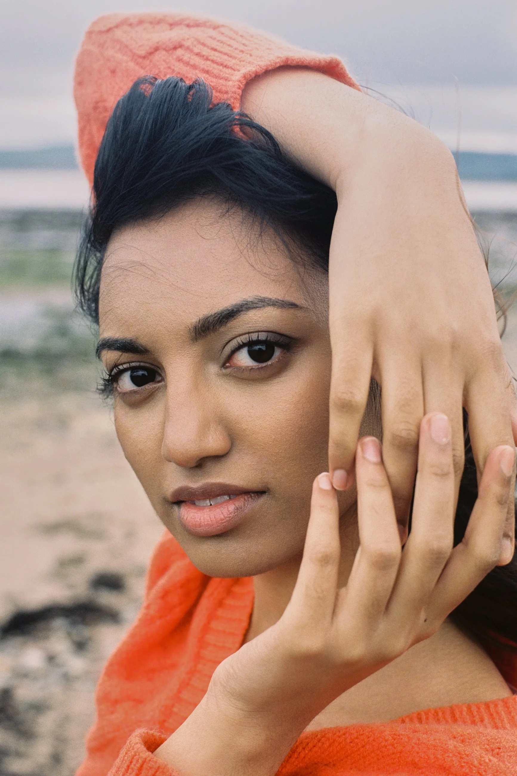 Close-up portrait of a young woman with dark hair on a beach, wearing an orange sweater, with her hand near her face and ocean in the background.