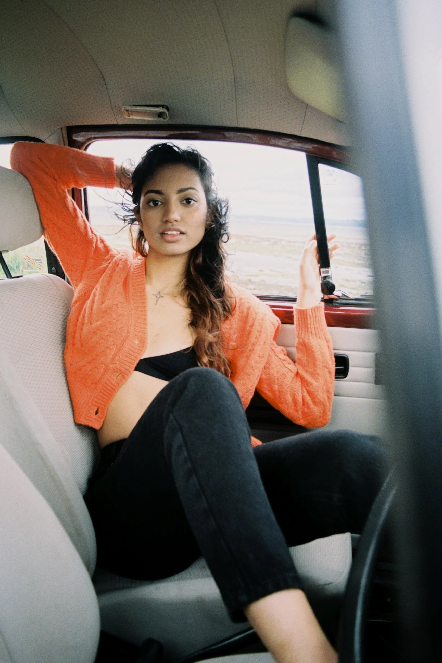 Young woman sitting in the passenger seat of a car, looking at the camera, with a casual pose, wearing a black top, an orange cardigan, and black pants, with a landscape view outside the window.