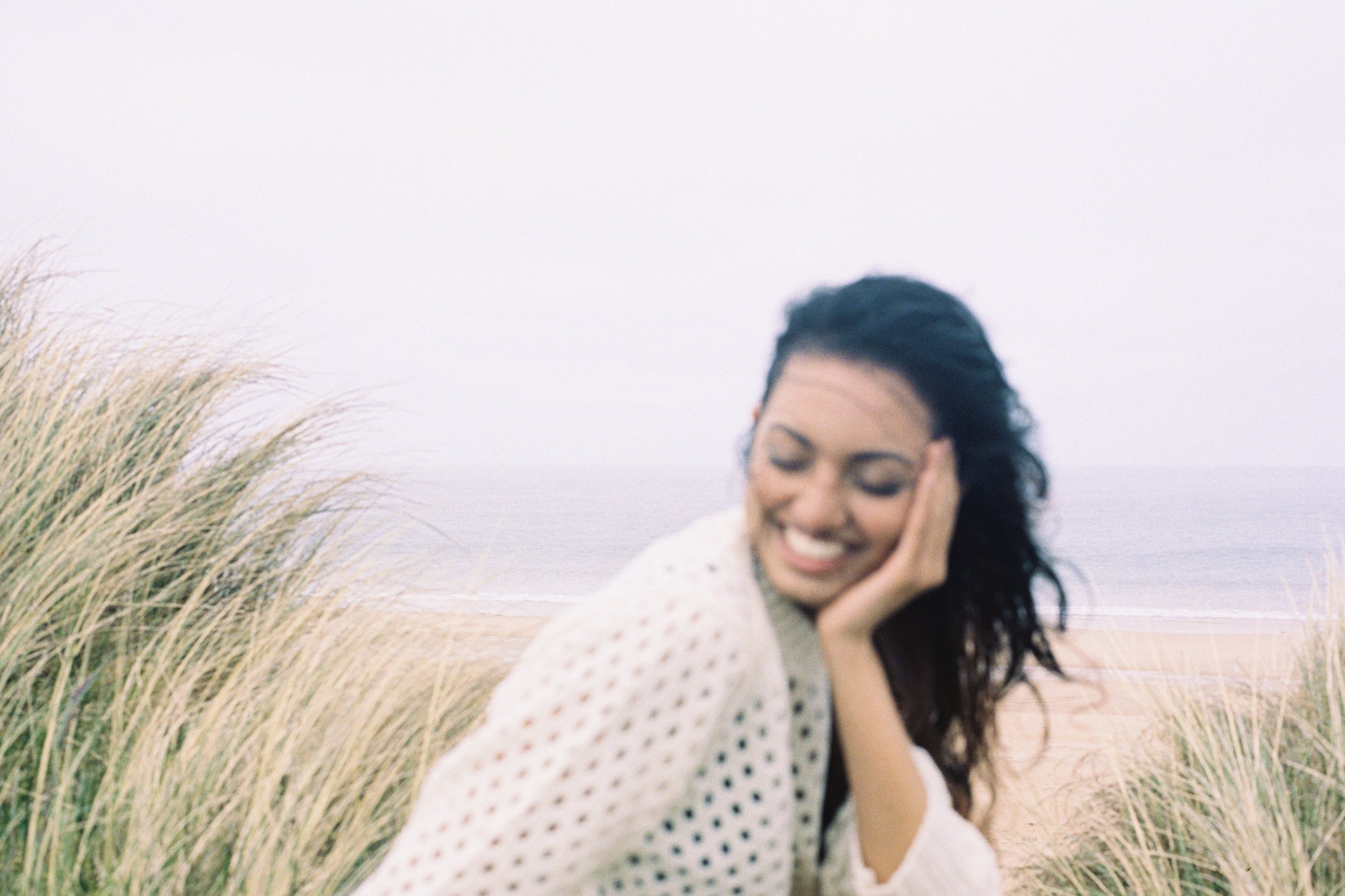 A woman smiling with her eyes closed, resting her face on her hand, standing between beach grass with the ocean in the background on a cloudy day.