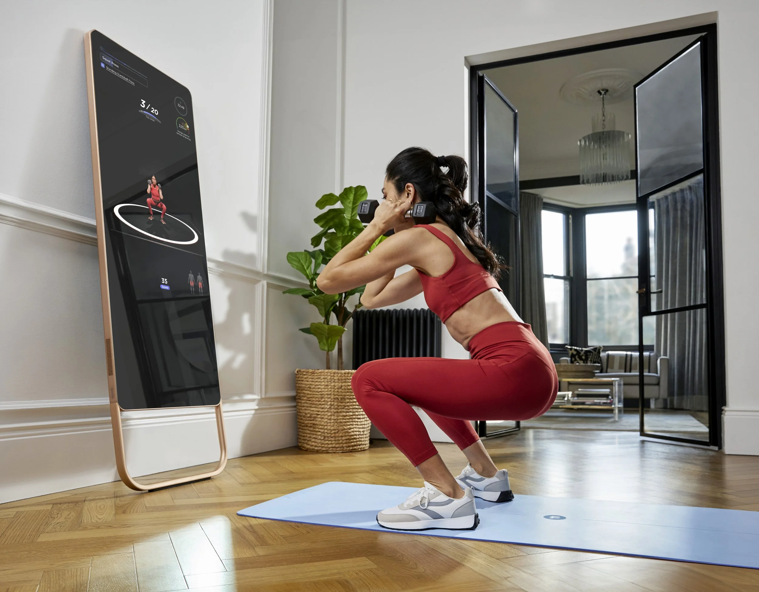 Woman in red workout clothes squatting on a blue yoga mat while holding dumbbells, following an on-screen fitness program on a large mirror-like device in a bright living room.