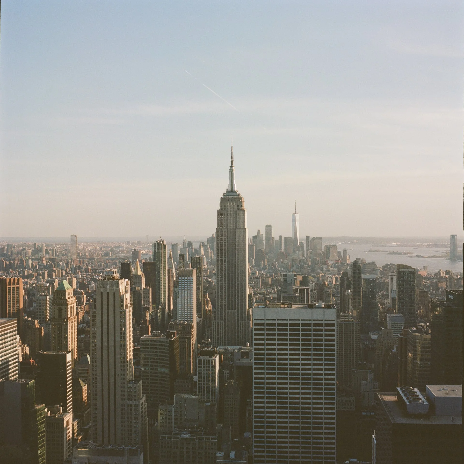 Cityscape view of New York City featuring the Empire State Building in the foreground and other skyscrapers with a clear sky and a faint airplane contrail.