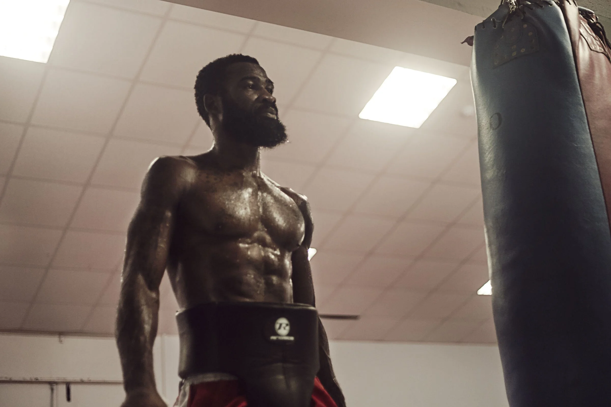A shirtless man with a beard and tattoos standing in a gym, looking at a punching bag.