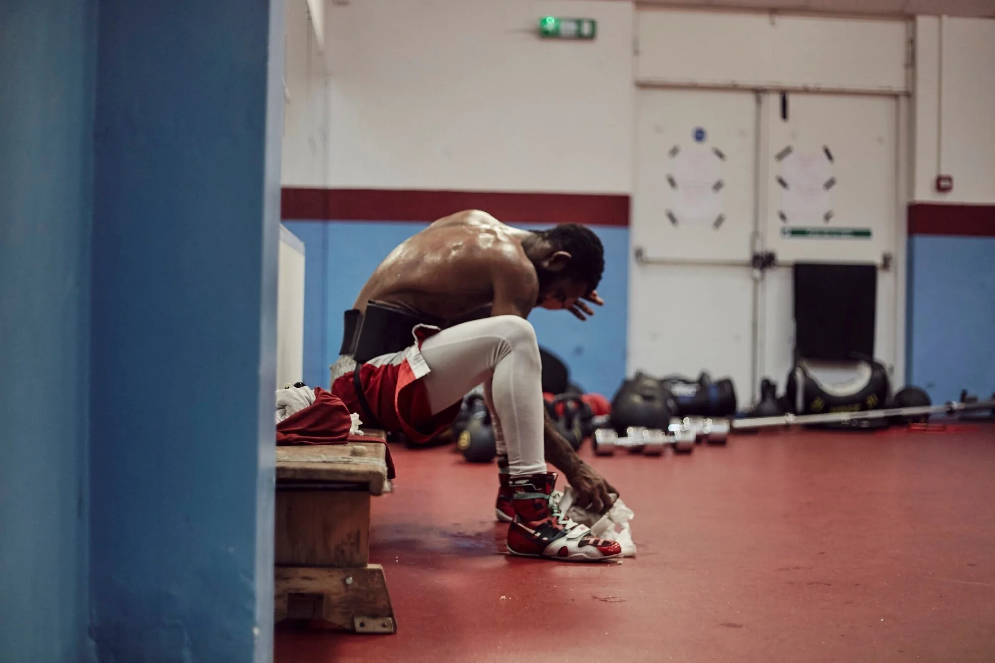 A male boxer sitting on a bench in a gym, putting on boxing gloves, with boxing equipment in the background.