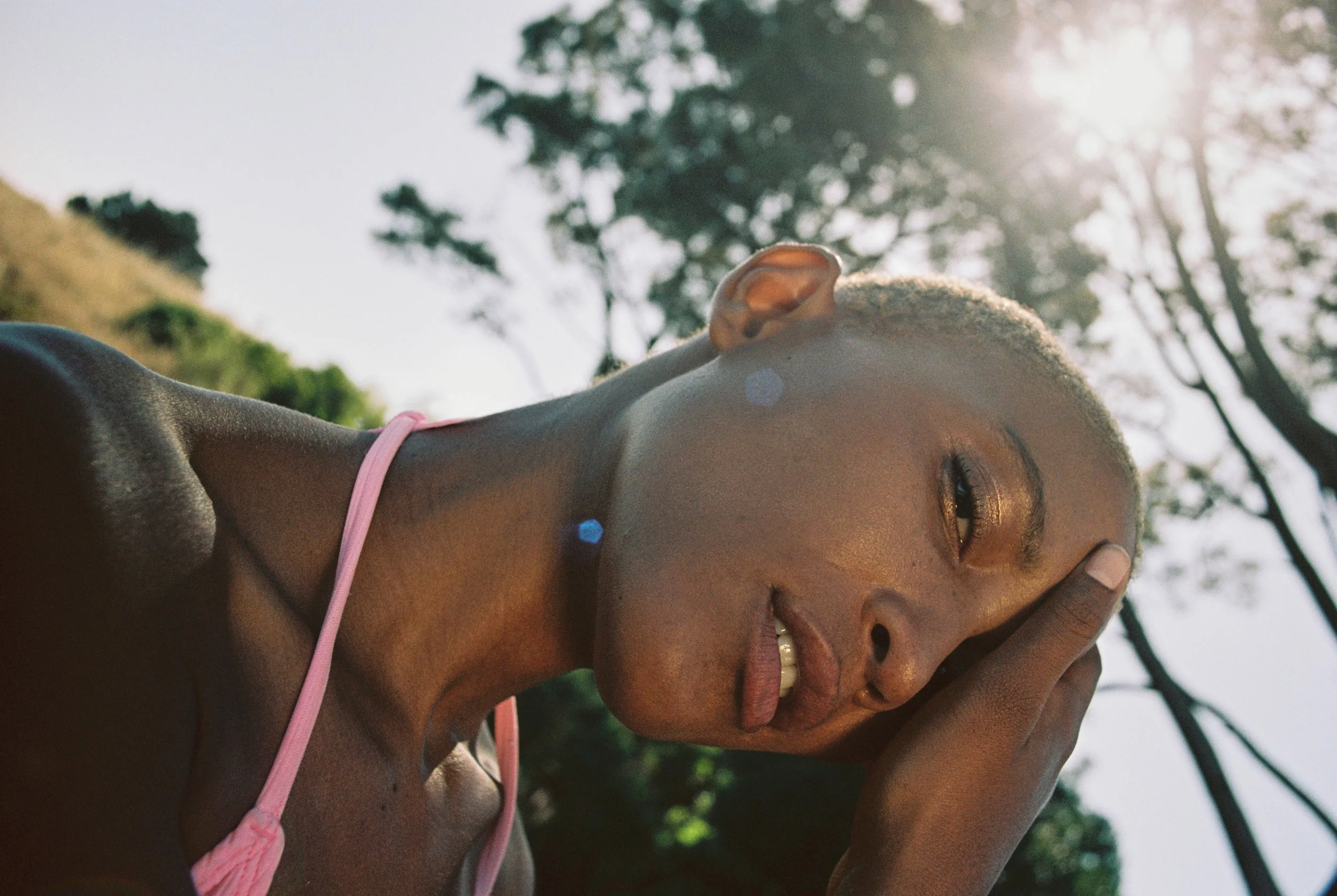 Close-up of a woman with short blonde hair, wearing a pink strap top, outdoors with trees and sunlight in the background.