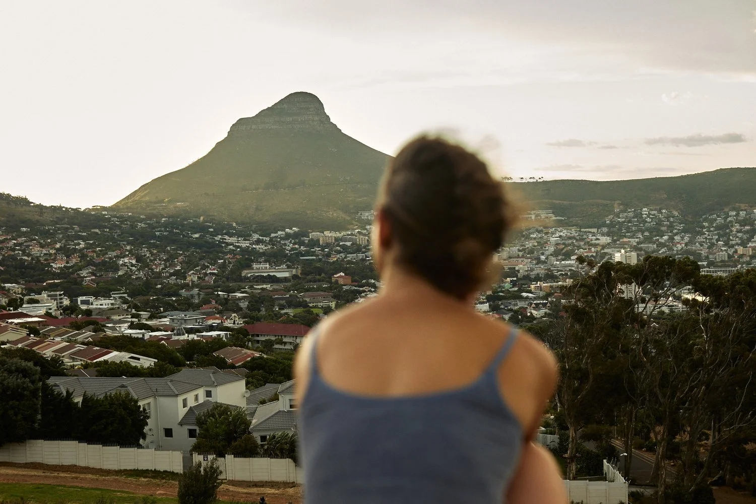 A woman with brown hair in a bun, wearing a blue tank top, looking at a cityscape with a mountain in the background, possibly during sunset.