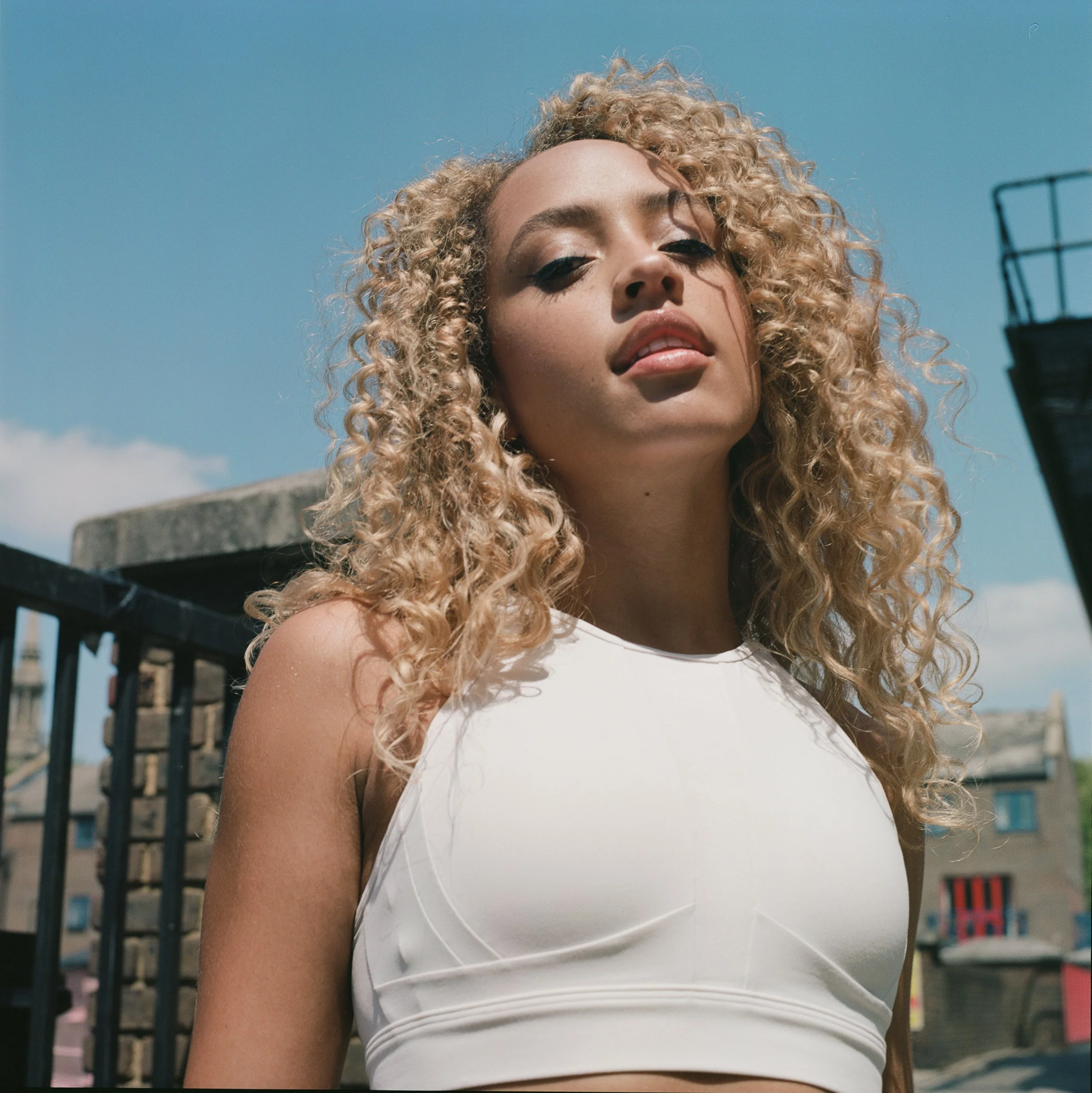 A woman with long curly blonde hair wearing a white sleeveless top, looking slightly down at the camera with a confident expression, outdoors with blue sky and buildings in the background.