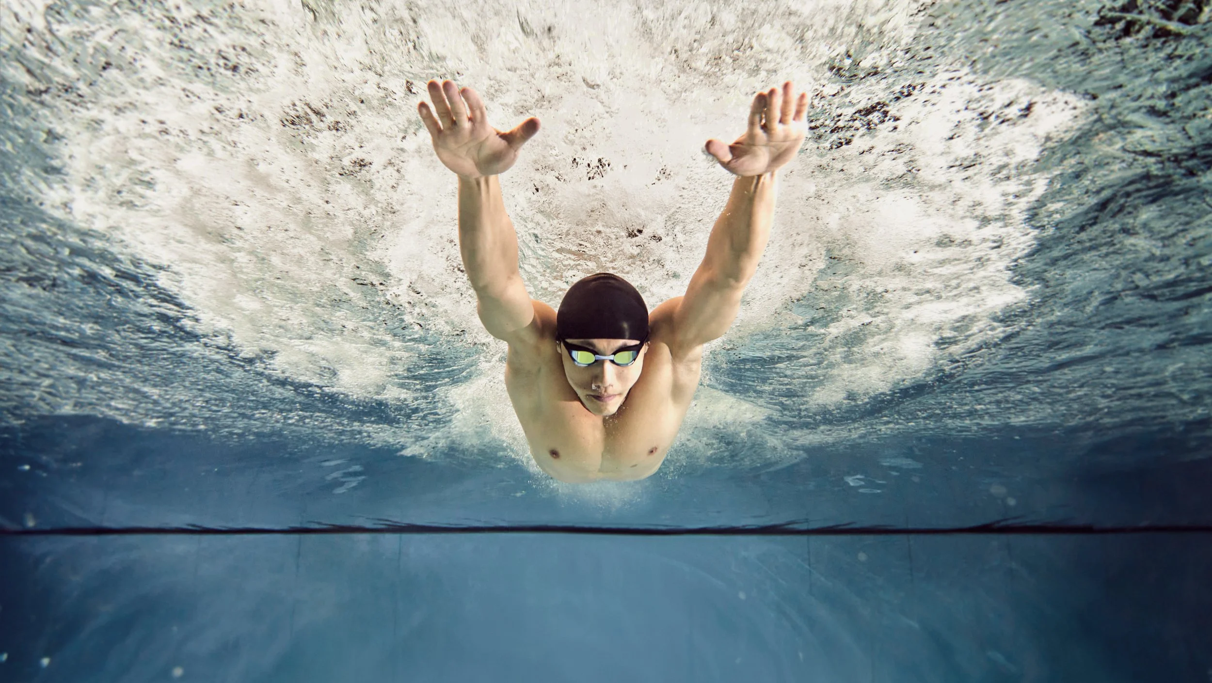 Swimmer in black cap and goggles swimming underwater, viewed from below.