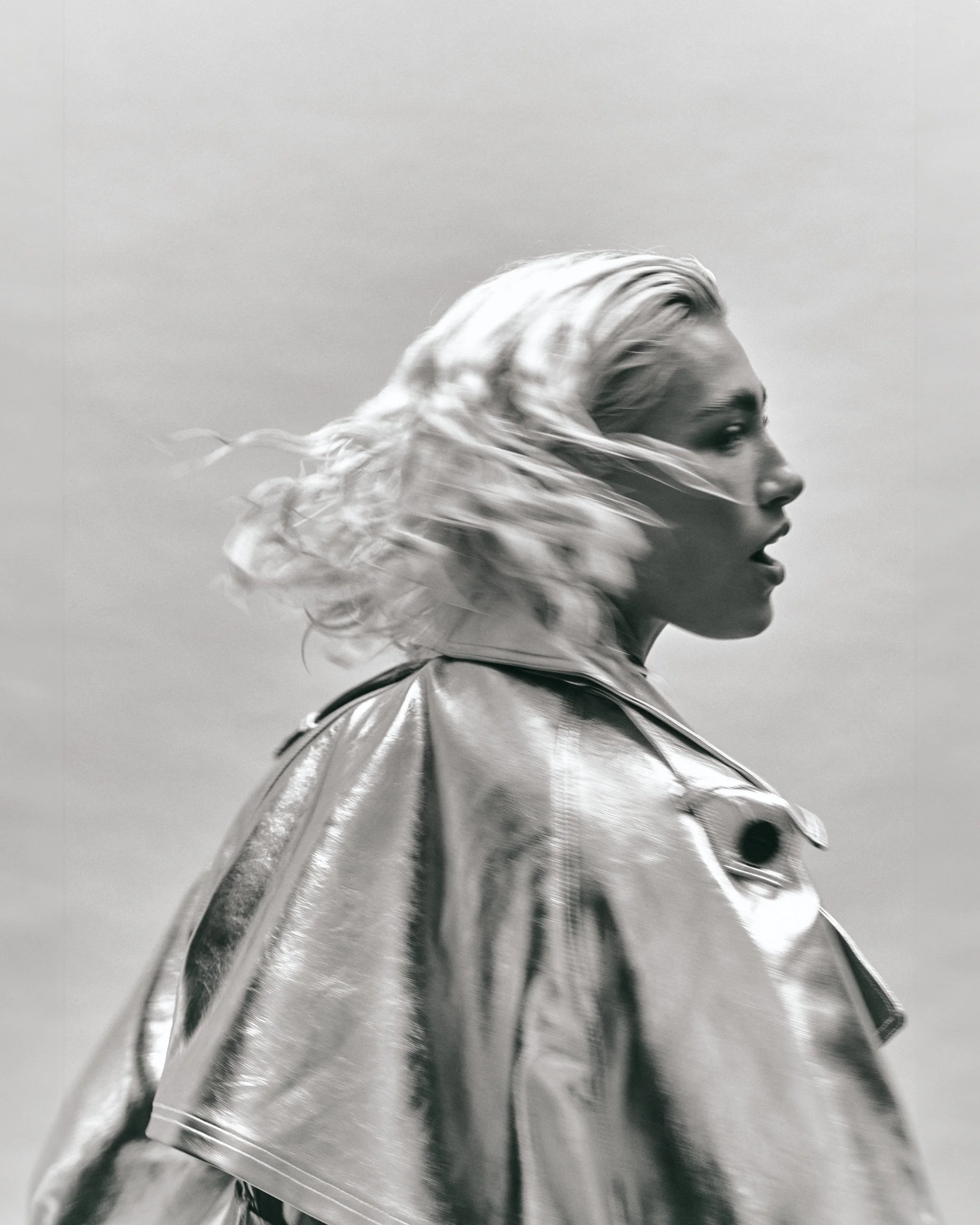 Black and white photo of a woman with blonde hair blowing in the wind, wearing a shiny metallic jacket, with a neutral background.