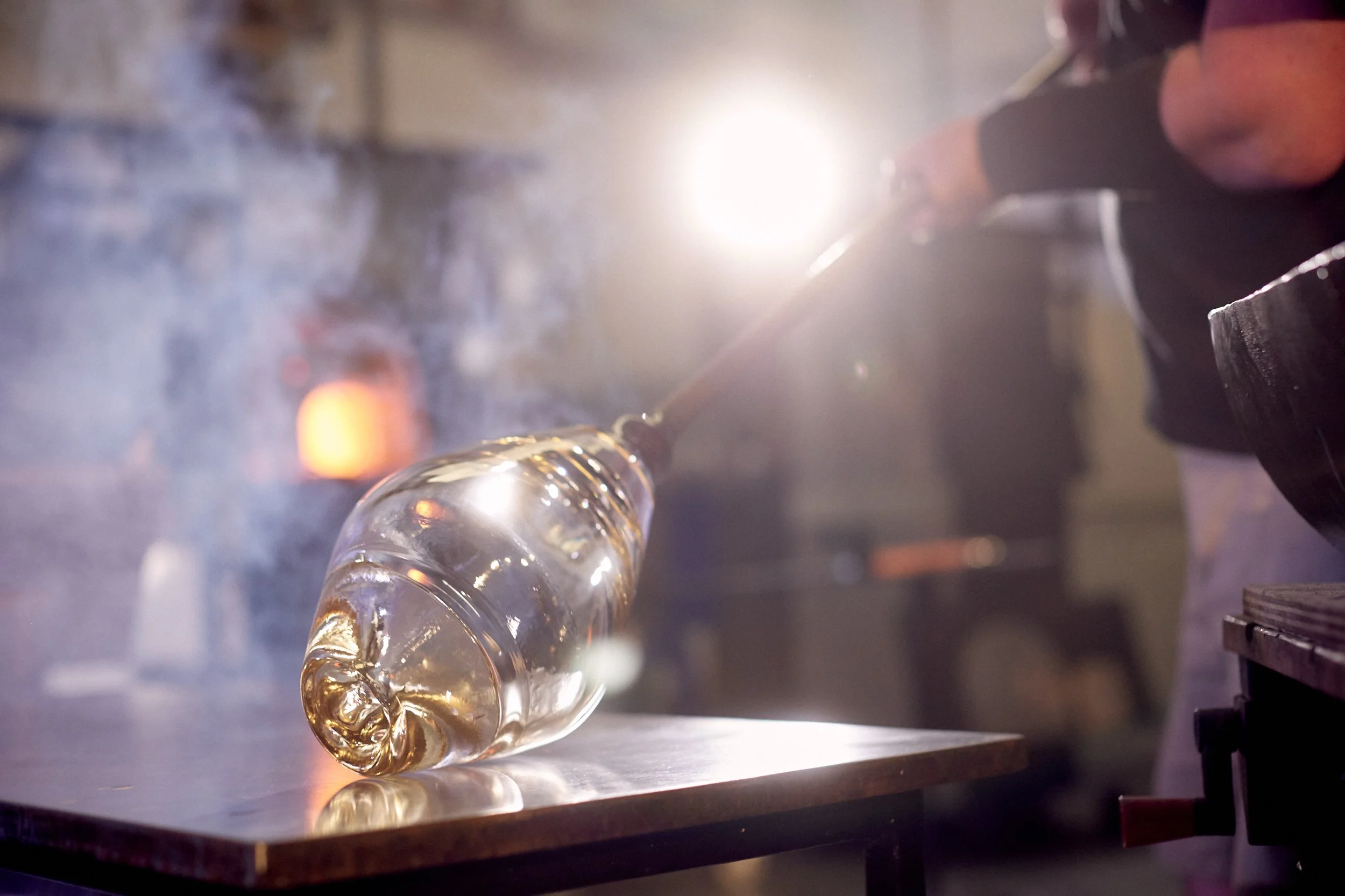 A glassblower using a blowtorch to shape a glass piece in a workshop with bright light and smoke.