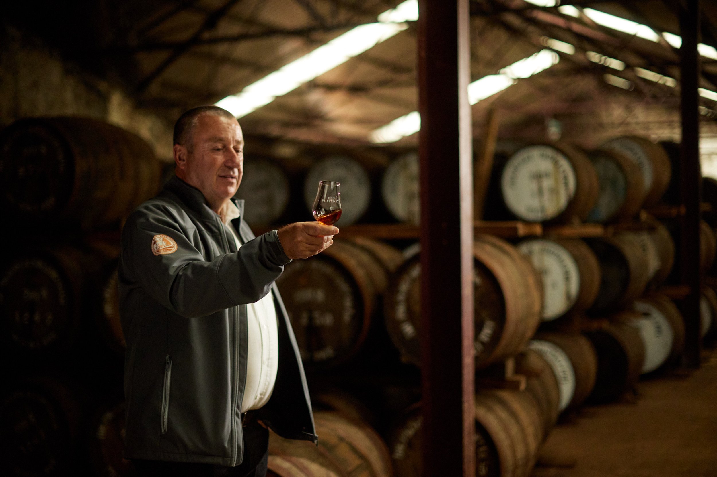A man in a gray jacket holding a glass of whiskey inside a distillery with wooden barrels stacked on shelves.