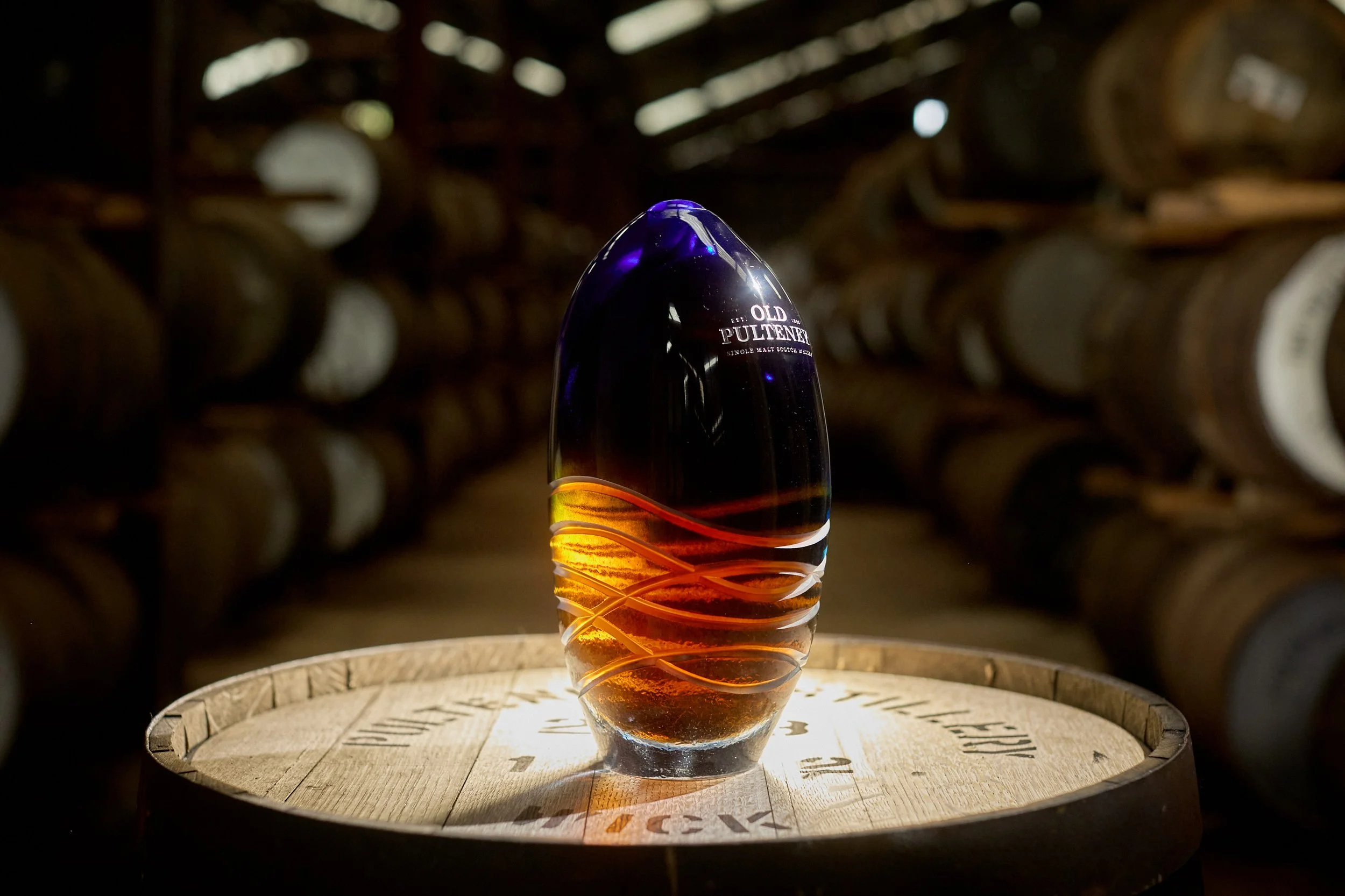 A glass sculpture of single malt whisky with a dark purple at the top fading to amber at the bottom, standing on a wooden barrel in a dimly lit storage room filled with stacked barrels.