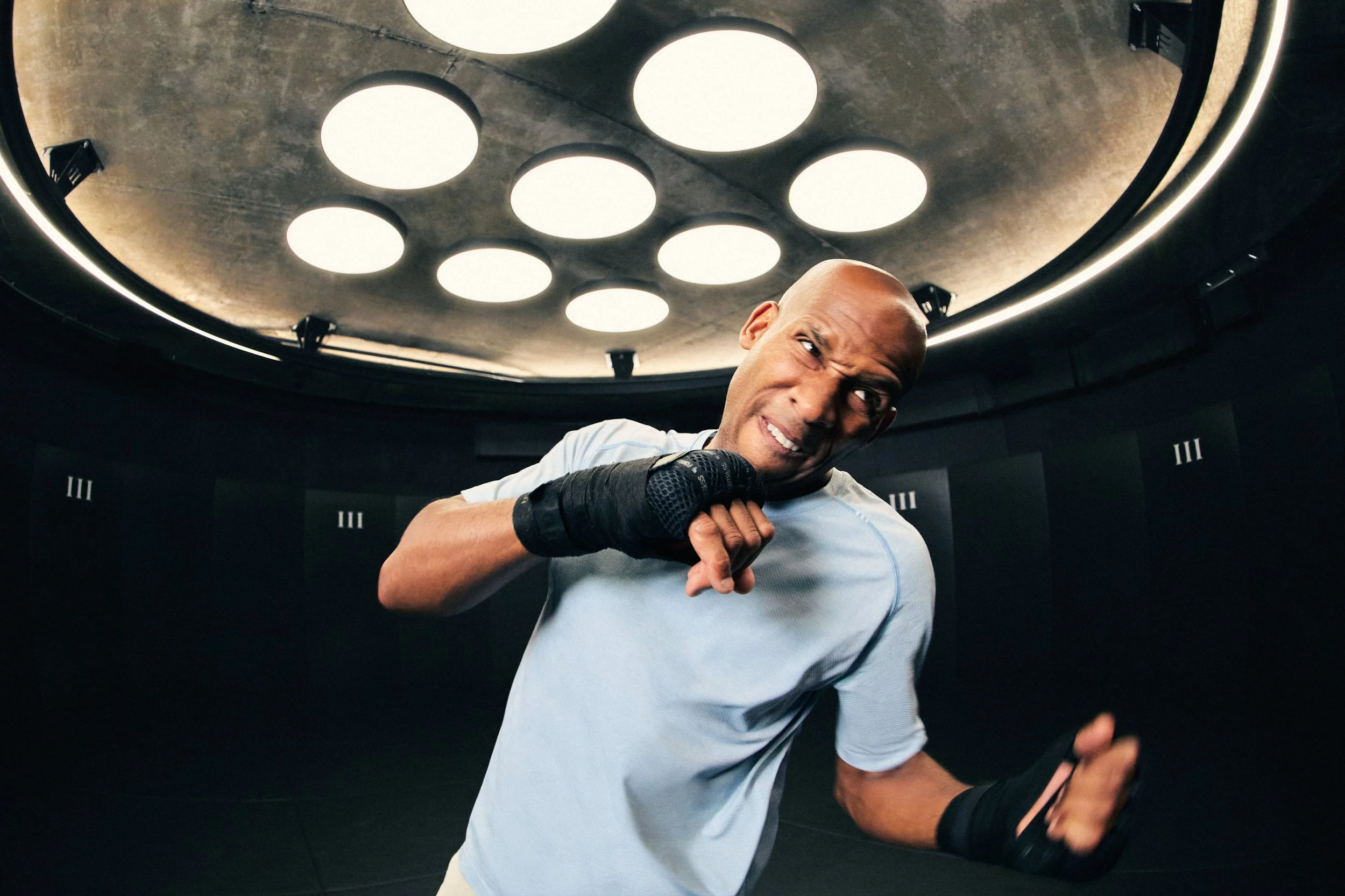 A man in boxing gear, wearing gloves, is striking a pose in a gym with lockers behind him and circular lights on the ceiling.