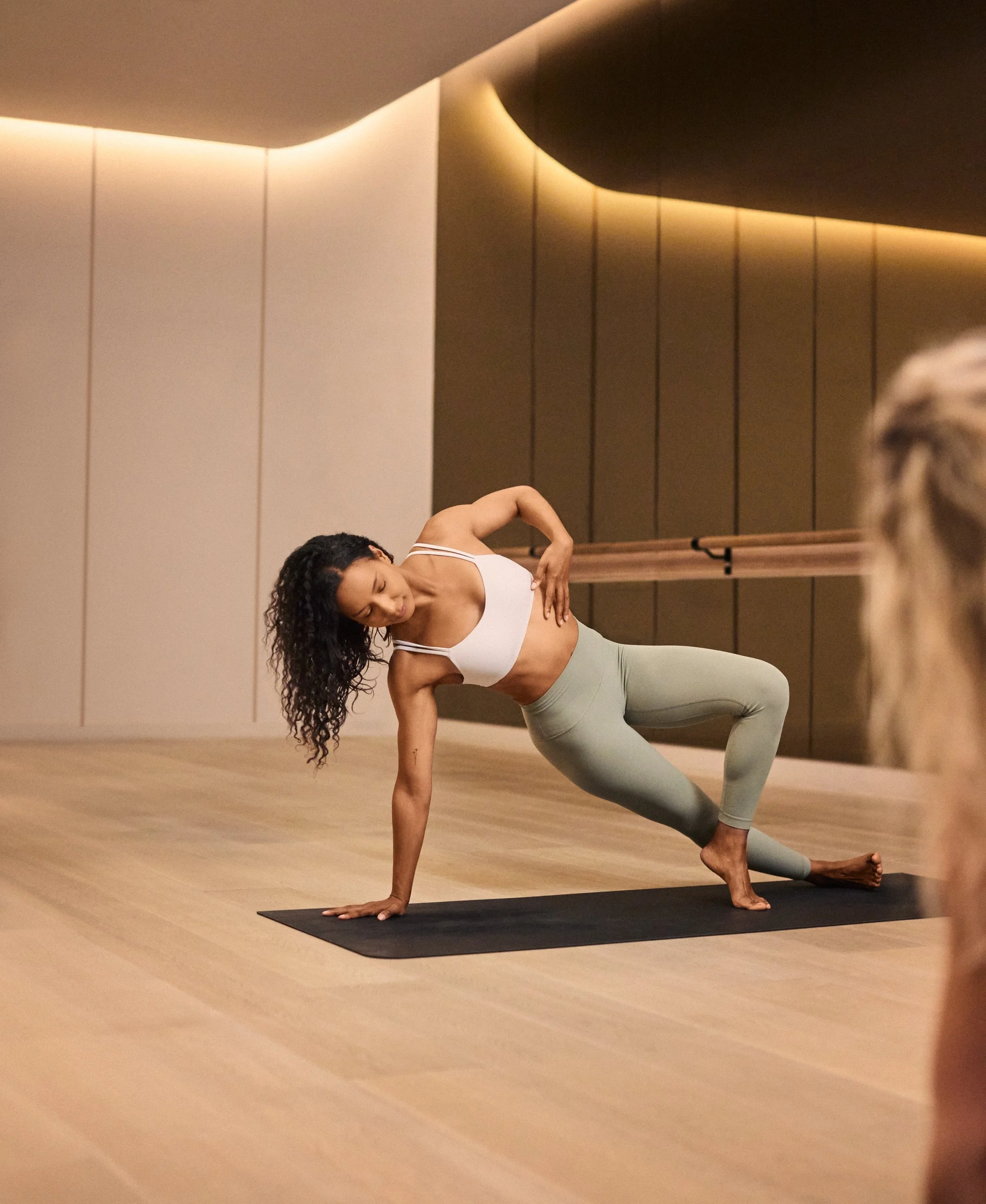 A woman with curly hair practicing yoga in a studio, doing a side plank pose on a black yoga mat.