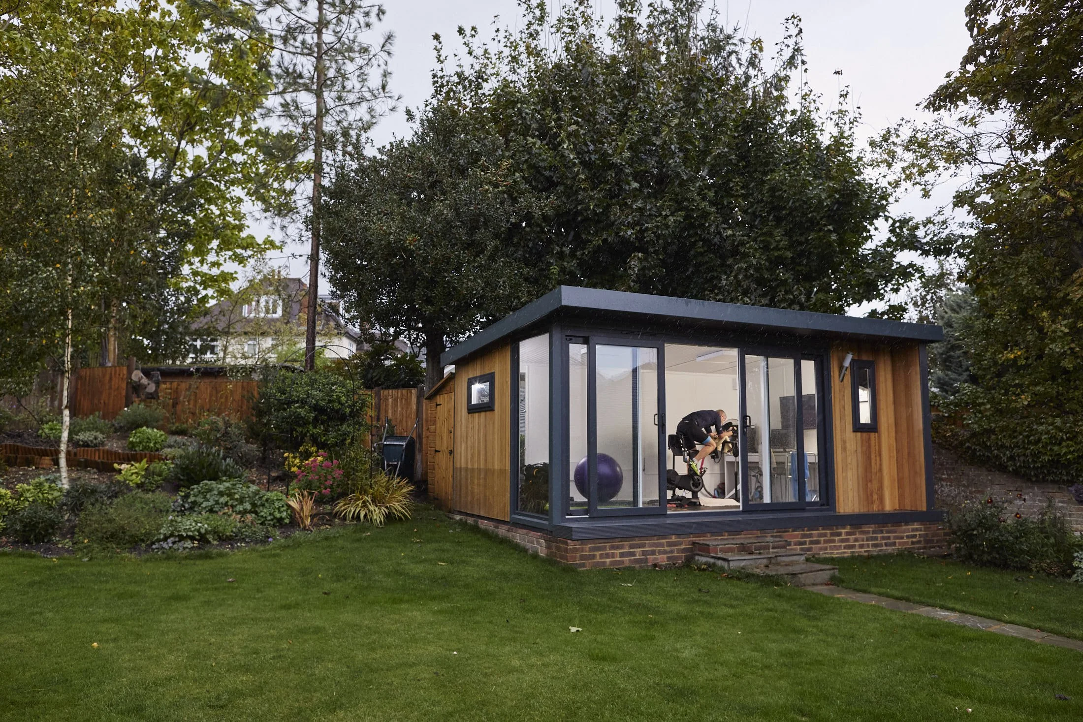 A backyard with a wooden and glass gym shed, a person exercising on a stationary bike inside, green grass, trees, and a garden with plants and flowers.