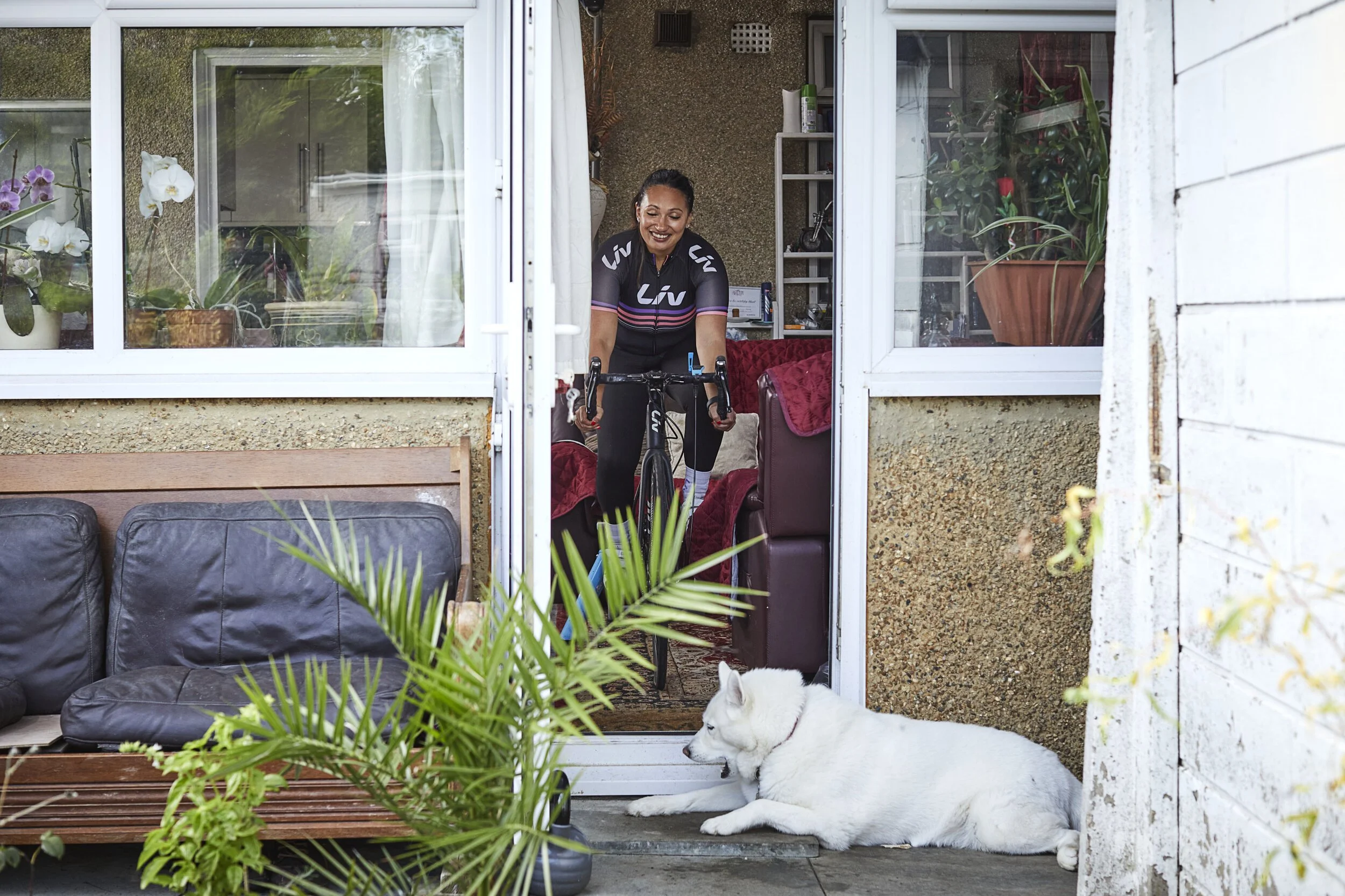 A woman in cycling gear is inside her home, smiling and holding a bicycle, seen through a sliding glass door. A large white dog is lying on the patio just outside the door, and there are green plants and outdoor furniture on the patio.