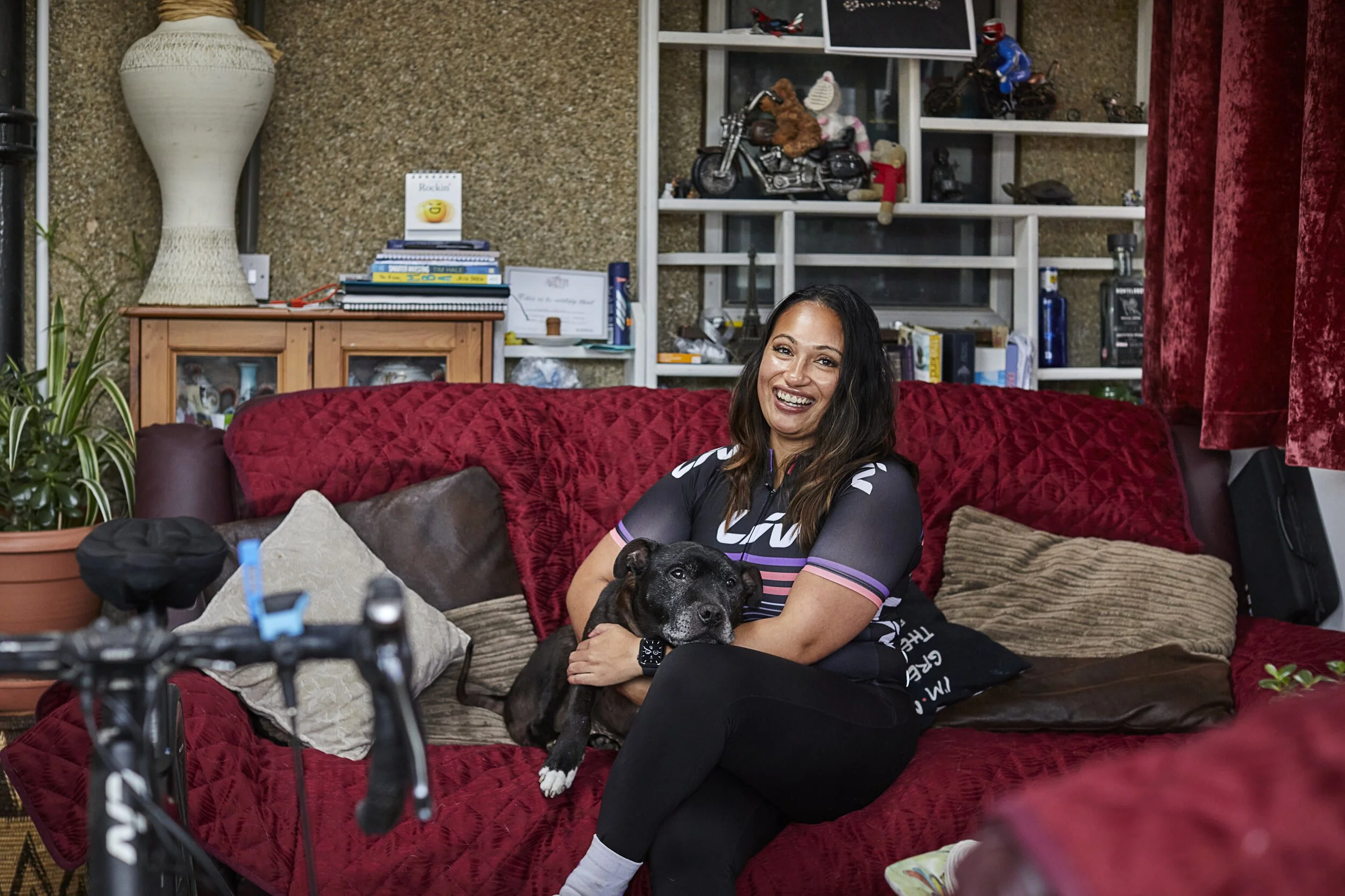 A woman sitting on a red couch, smiling, holding a black dog in her lap inside a cozy living room.