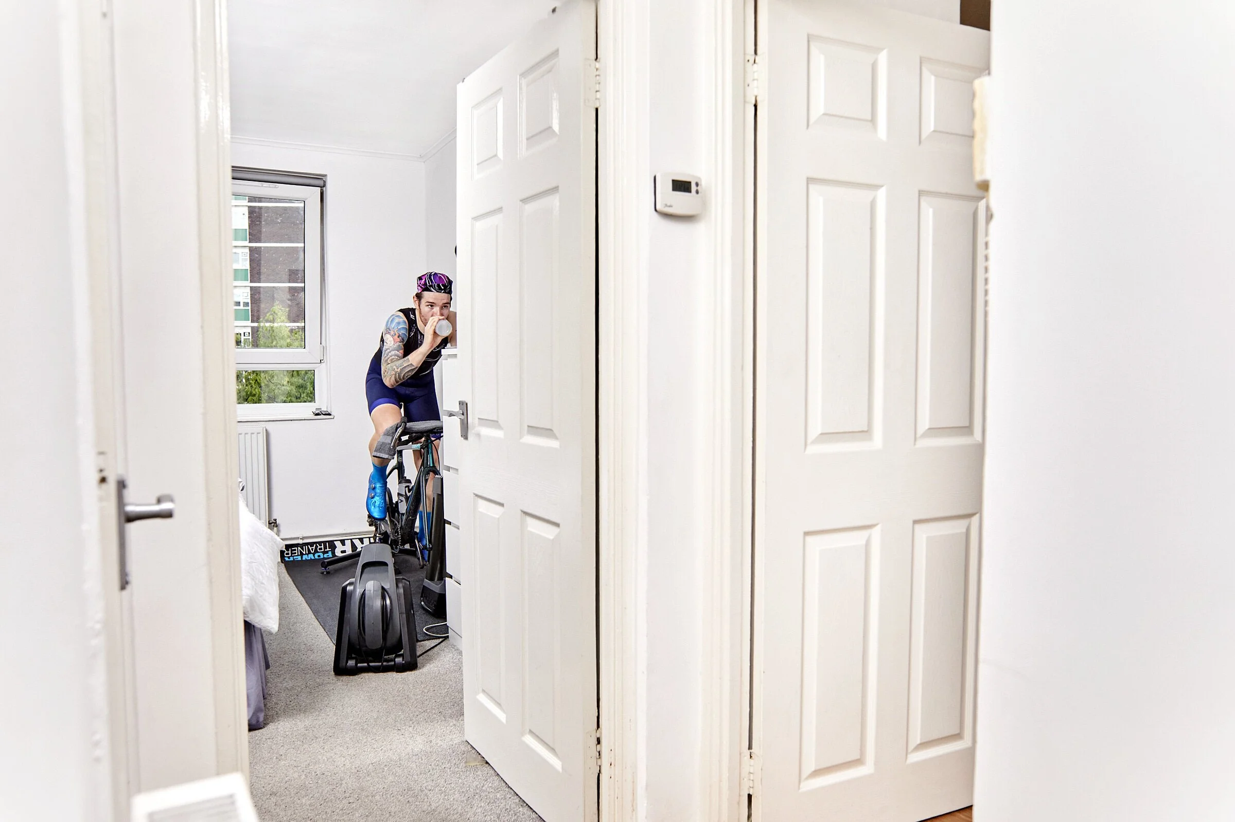 Person with tattoos on arms, wearing a purple and blue cycling outfit, drinking water while riding a stationary bike in a small, bright bedroom with white walls and a window.