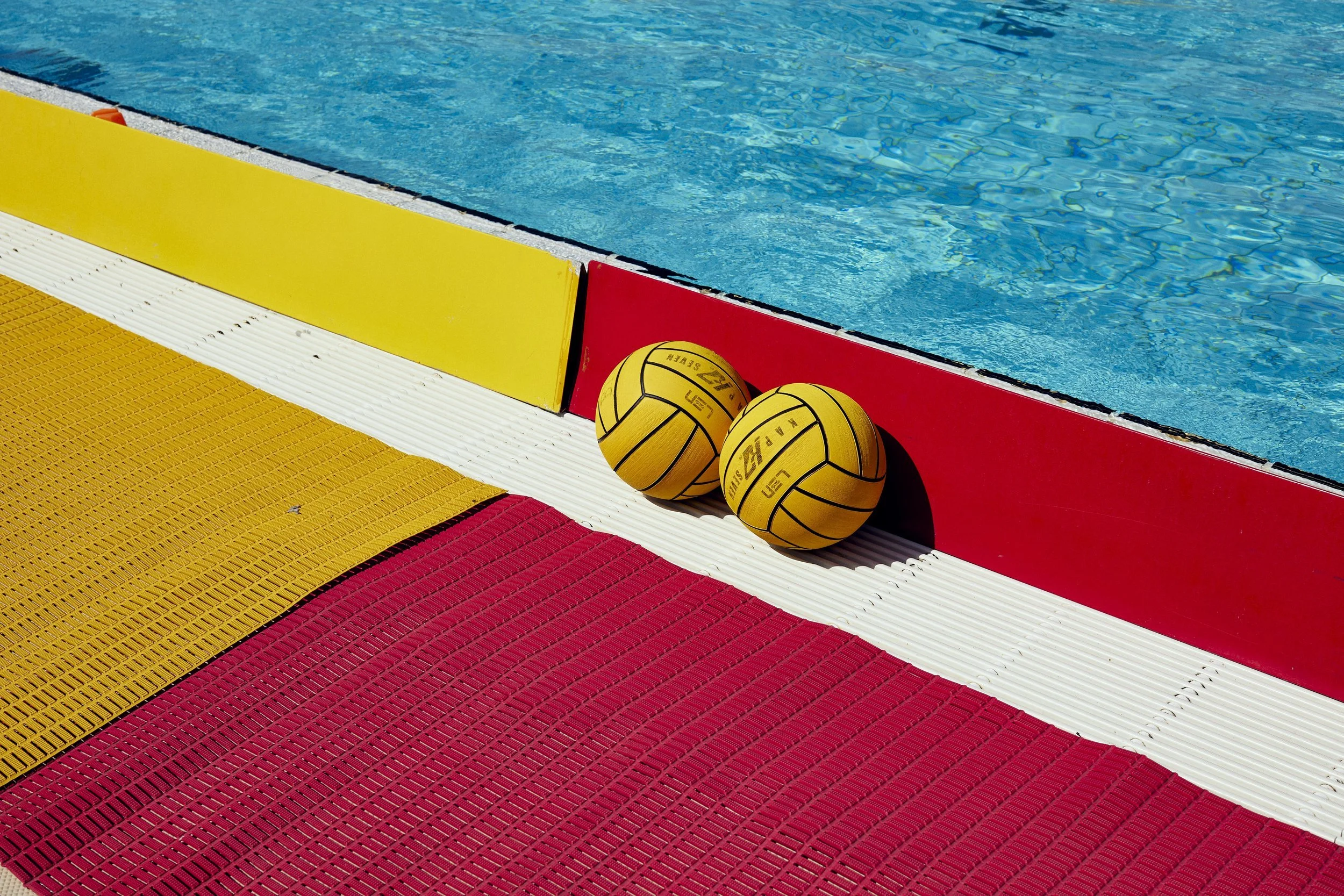 Two yellow volleyballs resting against the red and yellow poolside edging near a swimming pool.