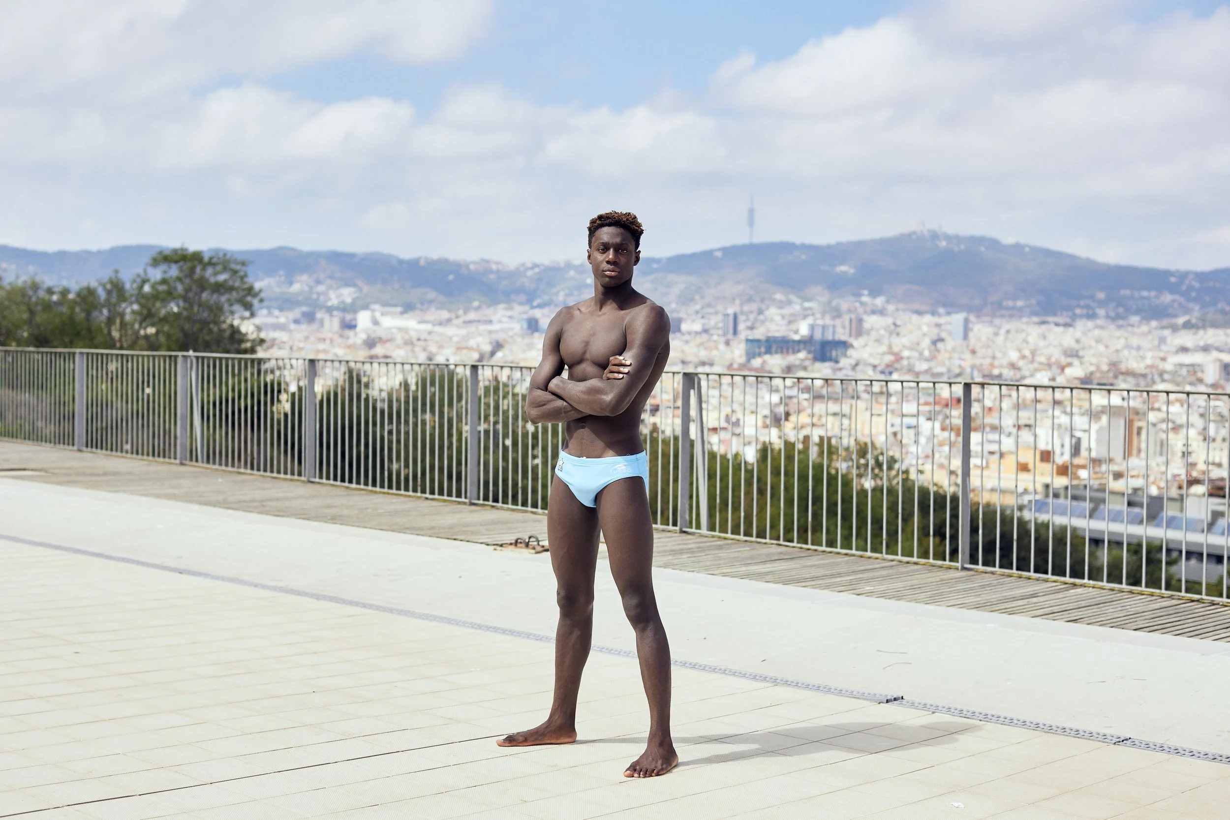A young man stands shirtless on a rooftop, wearing light blue swimwear, with a cityscape and hills in the background.