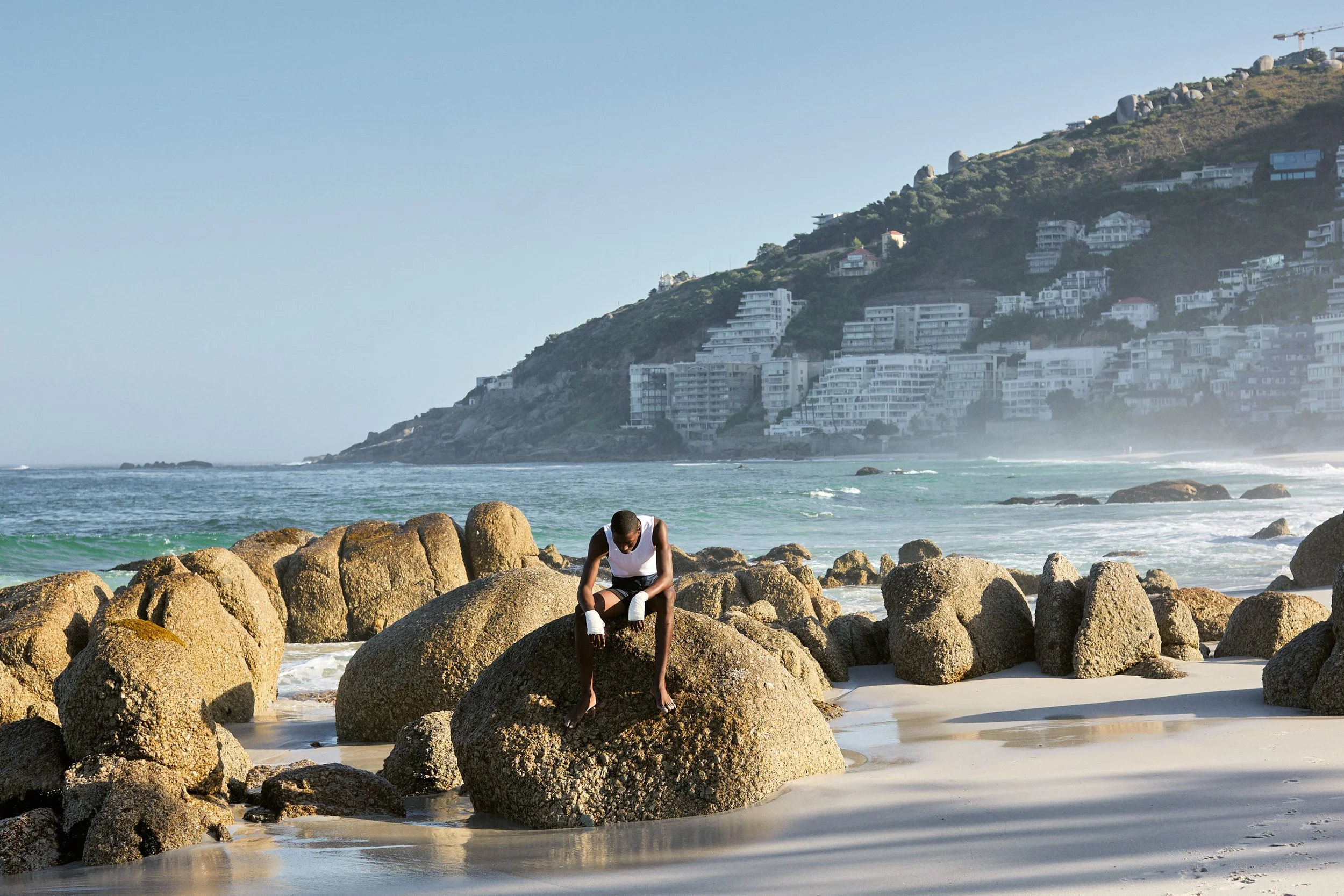 A person sitting on a large rock on a beach with other rocks, with ocean waves in the background and buildings on a hillside.