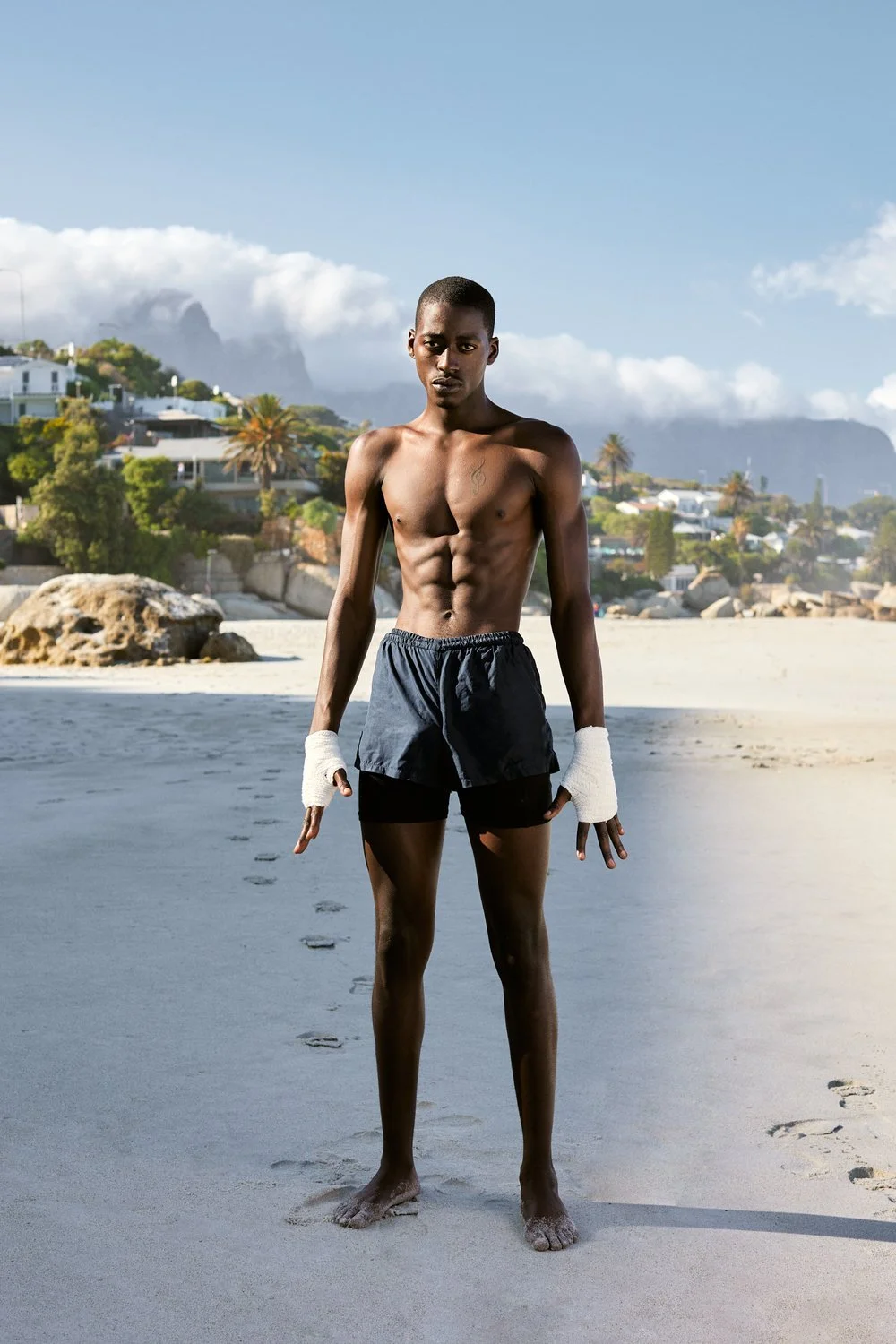 A shirtless young man with athletic build and dark skin stands on a sandy beach, wearing black shorts and white hand wraps, with a background of houses, palm trees, rocks, and mountains under a partly cloudy sky.