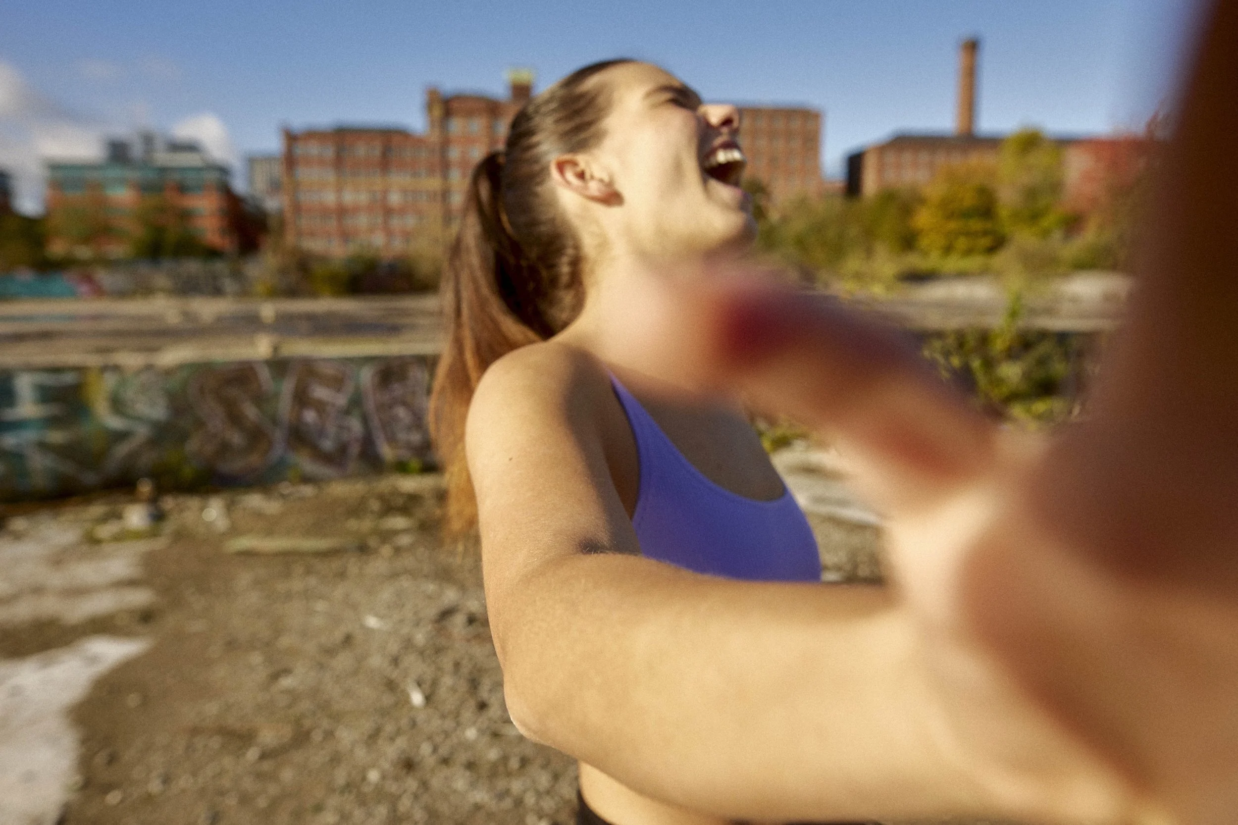 A young woman with long brown hair in a ponytail, wearing a purple tank top, laughing with her eyes closed, outside on a sunny day with a cityscape background.