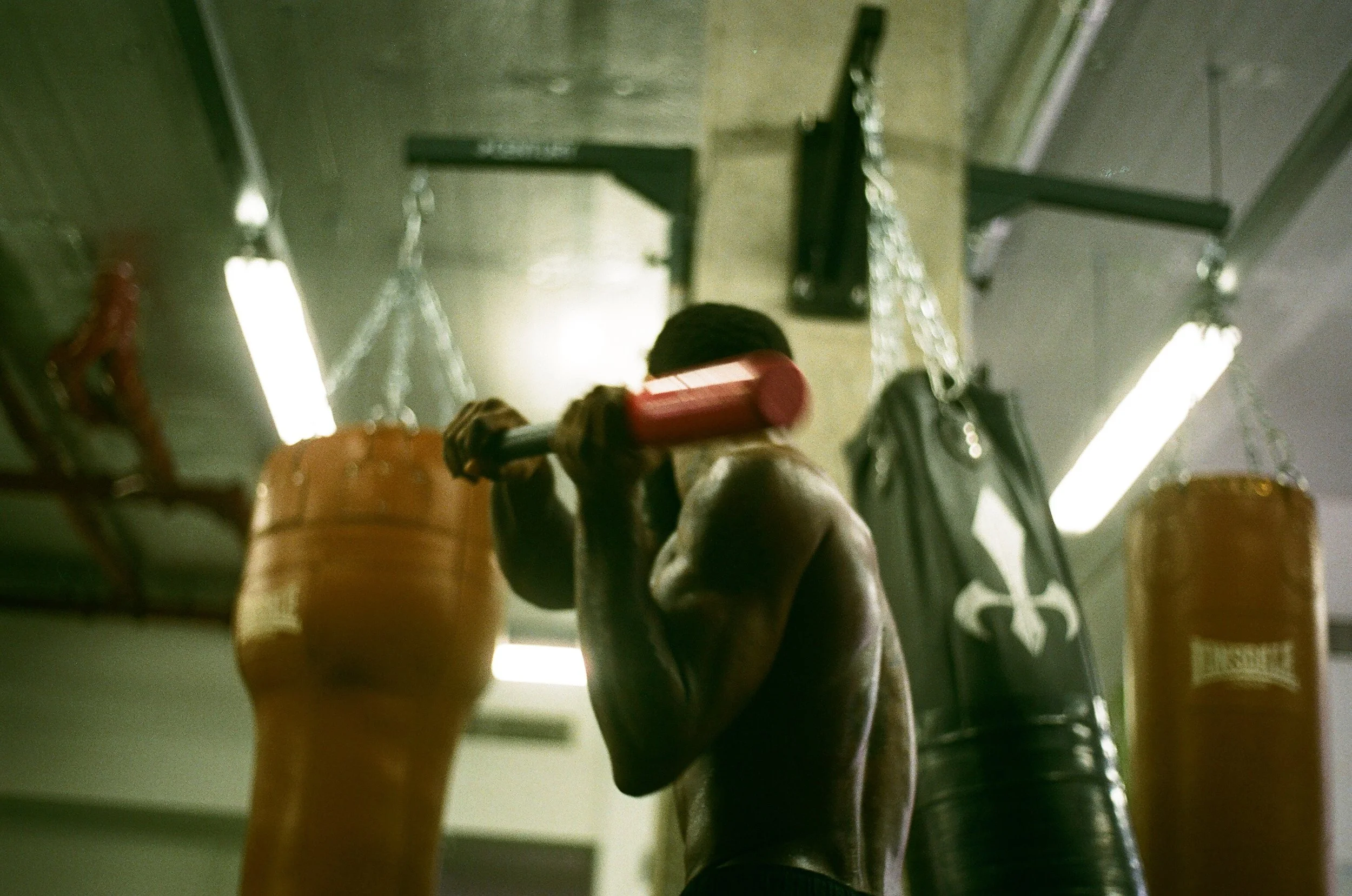 A person with a muscular build is training in a boxing gym, holding a red and black barbell on shoulders, facing a hanging punching bag.
