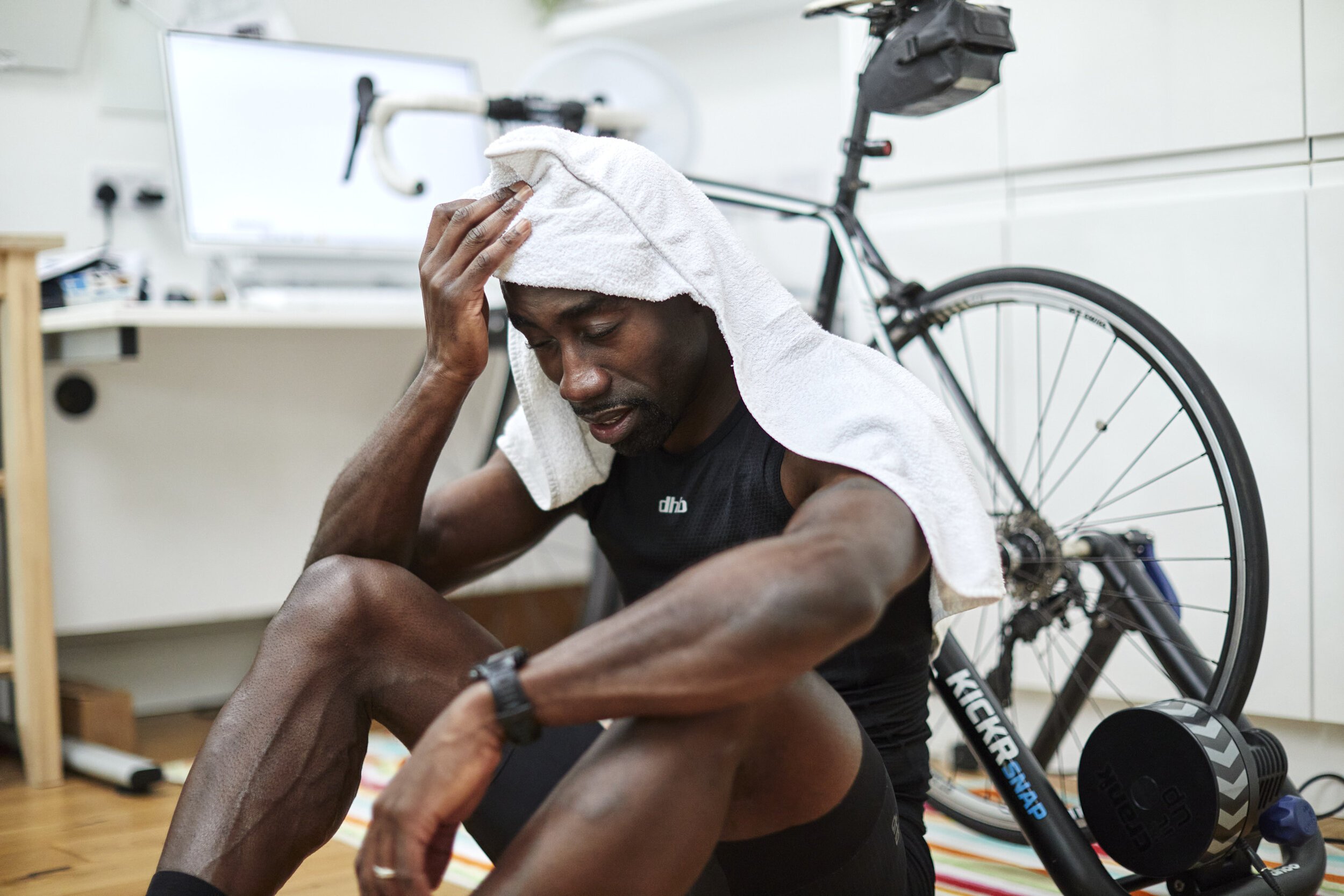 Man dressed in athletic gear sitting on the floor with towel on his head, holding his knee with an expression of fatigue or pain, in a room with a bicycle and a computer in the background.