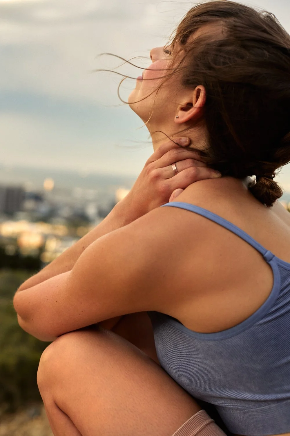 Woman in blue tank top sitting outdoors, holding her neck with windblown hair and cityscape in the background.